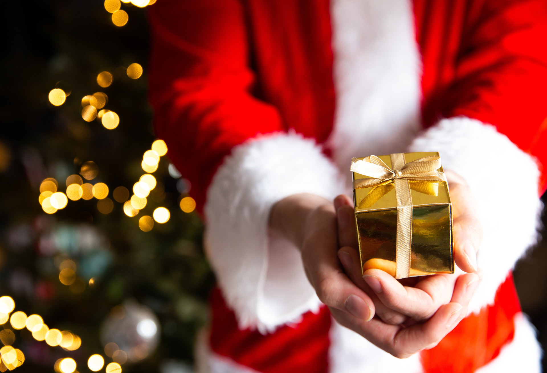 Man in Santa Claus costume near Christmas tree holding a golden gift box, Christmas present background with bokeh lights beauty