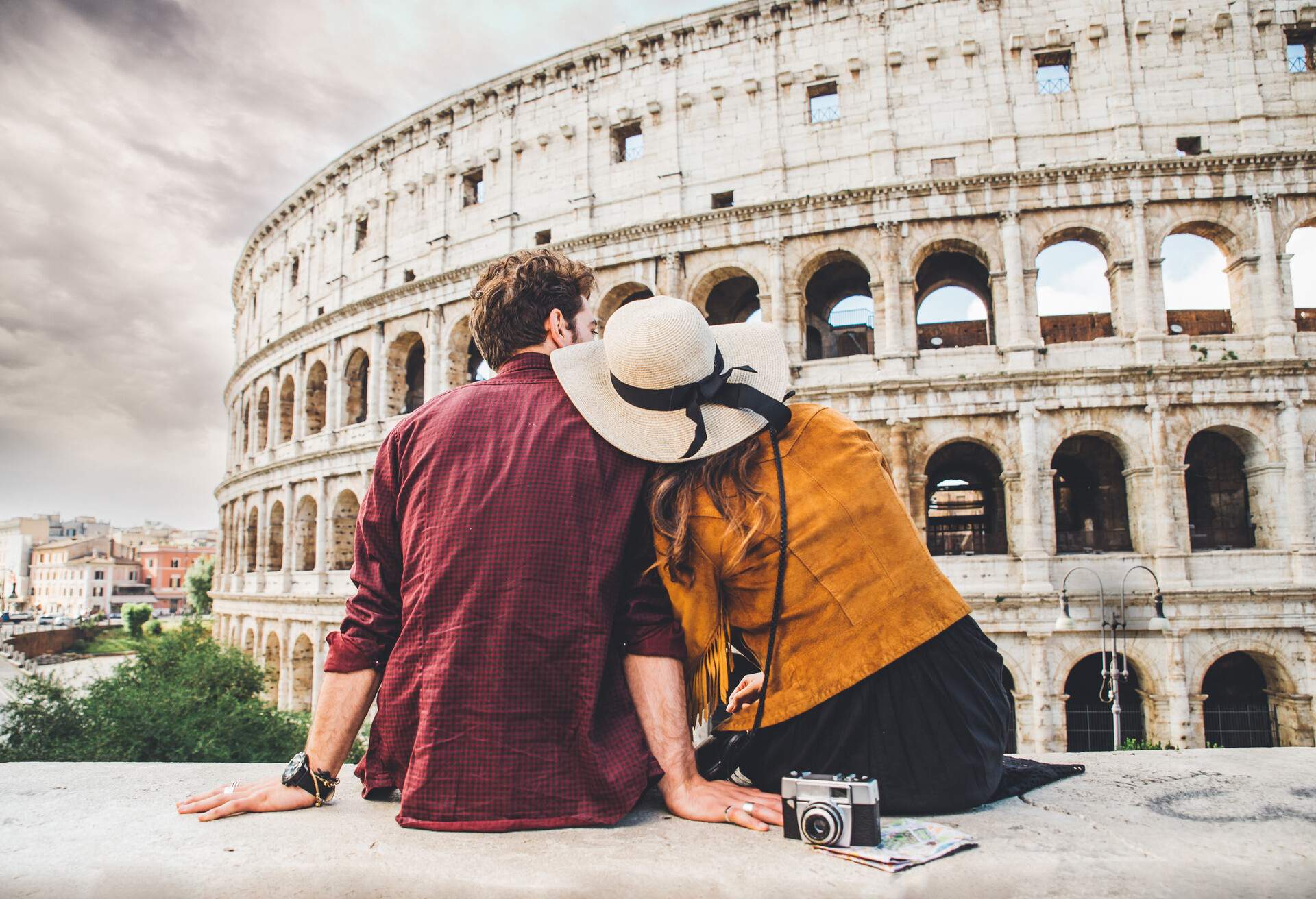 A woman in a wide-brimmed hat sits on a ledge beside a man while resting her head on his shoulder in front of Rome's Colosseum.