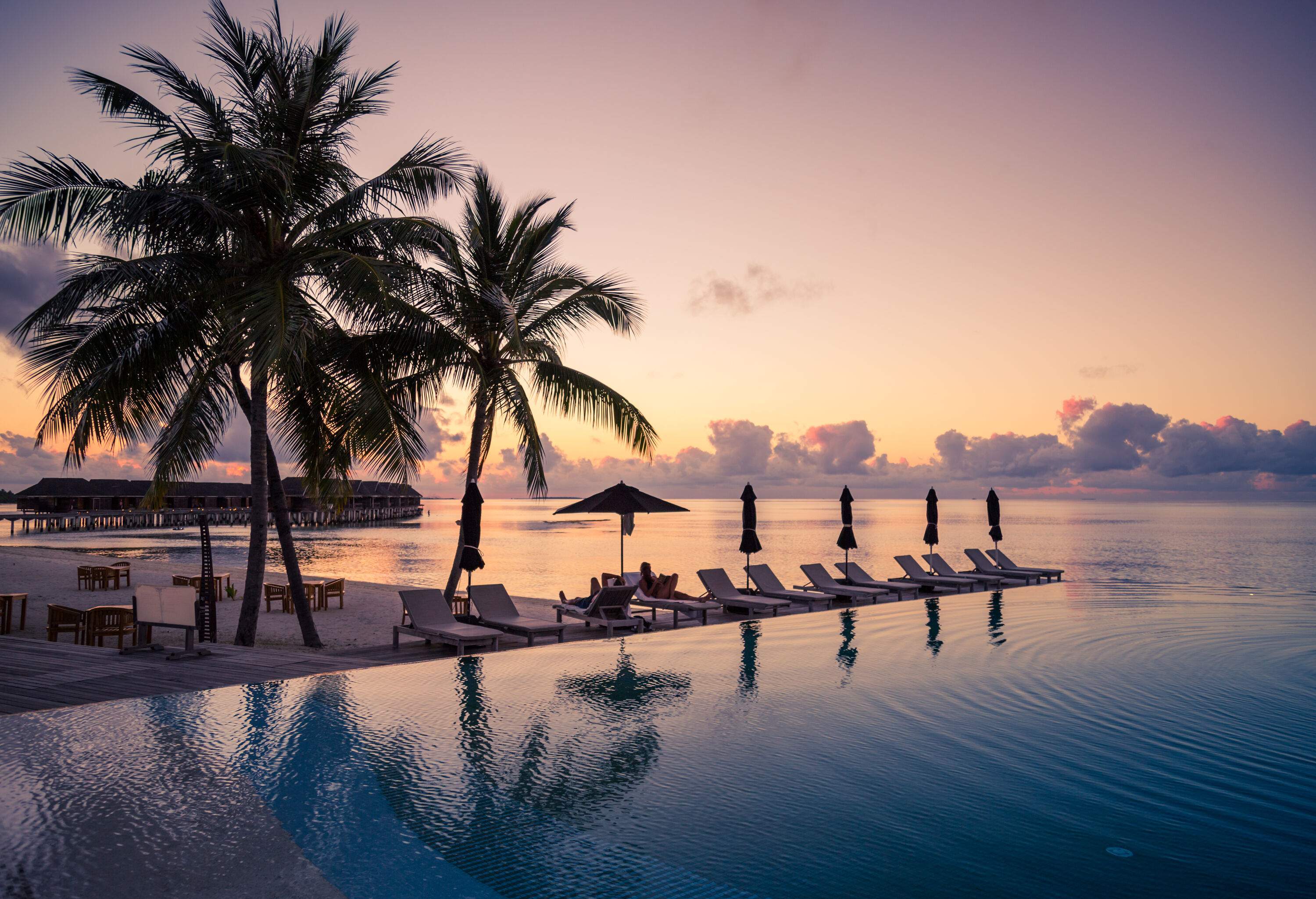 A couple relaxing on the sunbeds beside the dazzling infinity pool against the tranquil ocean on a scenic sunset.
