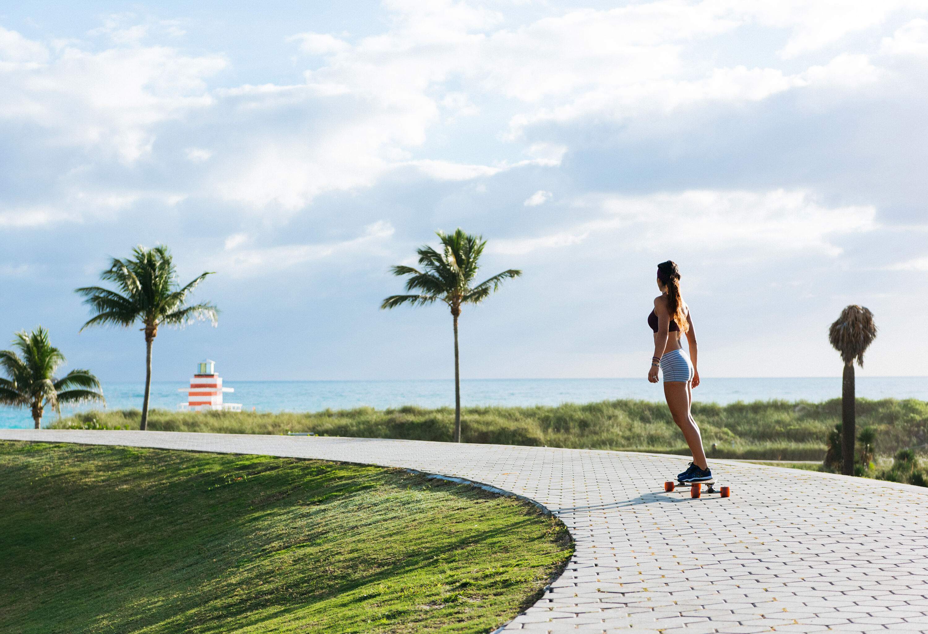 A woman skateboards down a curved concrete path on a grassy hill with vast views of the ocean.
