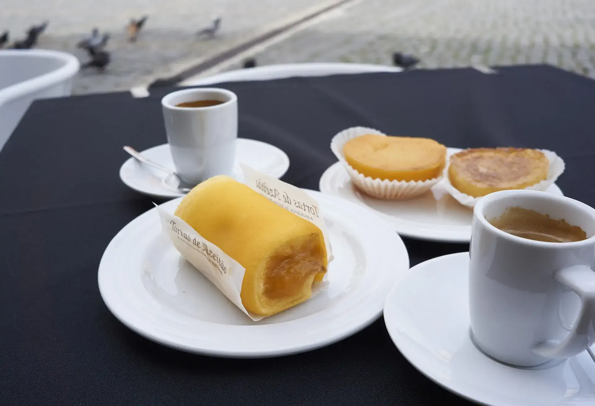 Torta de Azeitão on a white plate with coffees and other pastries on a black table cloth