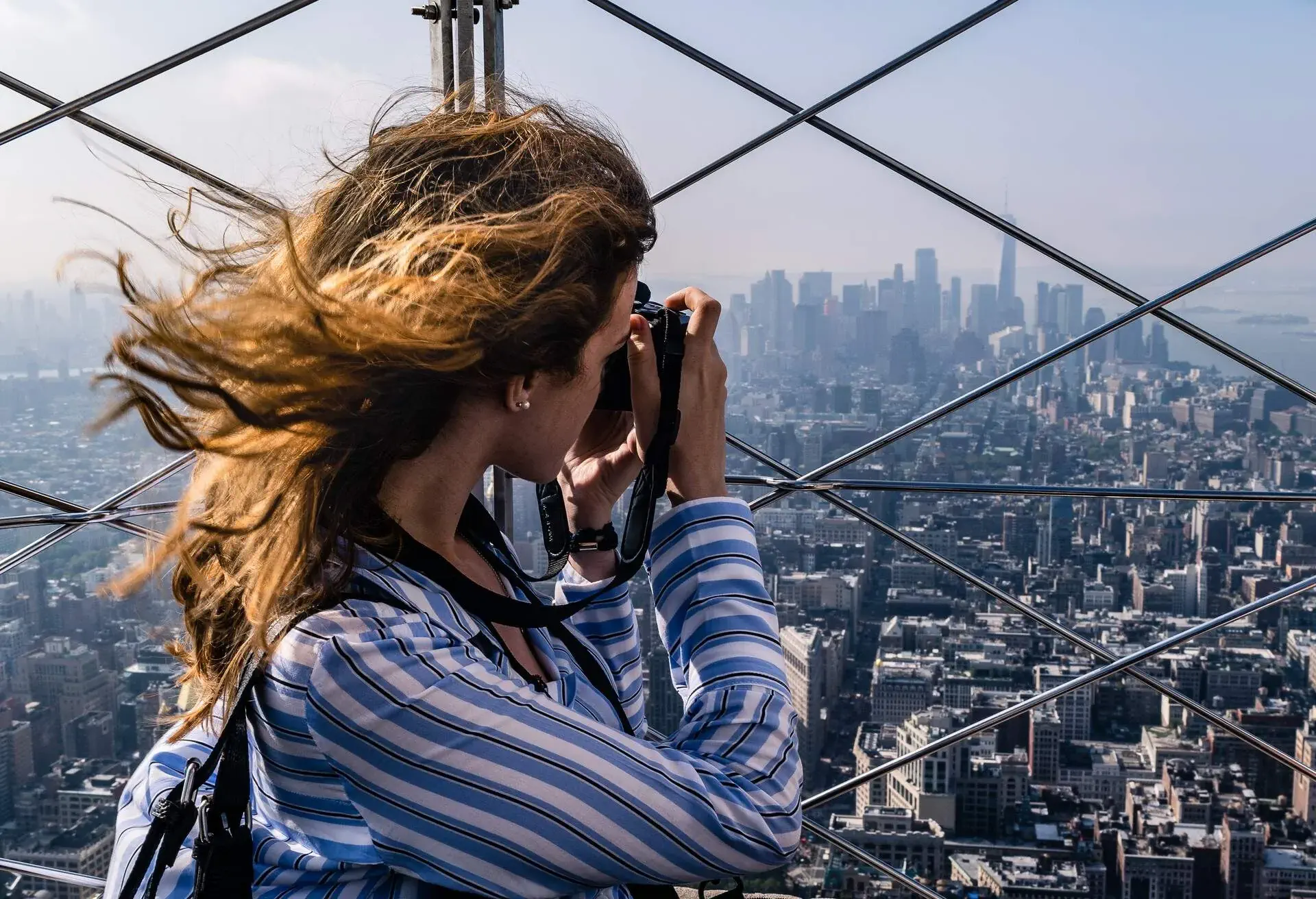 A brunette woman in a striped shirt photographs a cityscape from a fenced rooftop as her hair moves through the breeze.