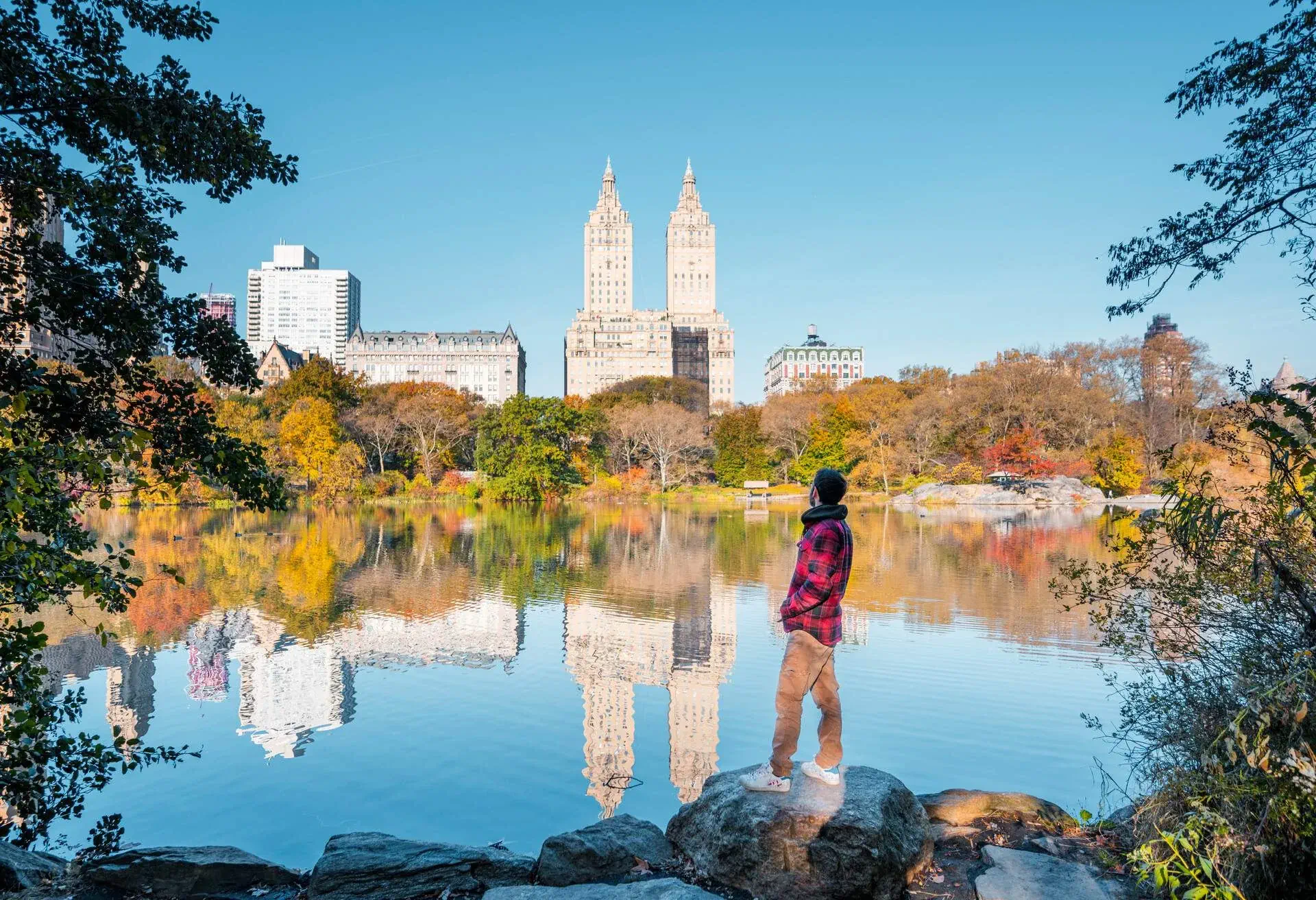 A man in a red flannel jacket standing on a rock by the pond in Central Park.