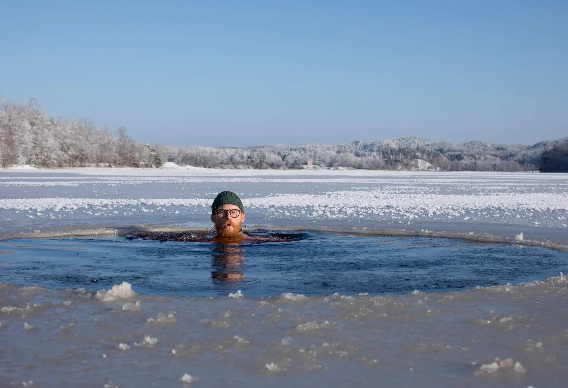 Bearded man wearing glasses and a wool hat swimming in an ice hole in frozen lake on a sunny winter day