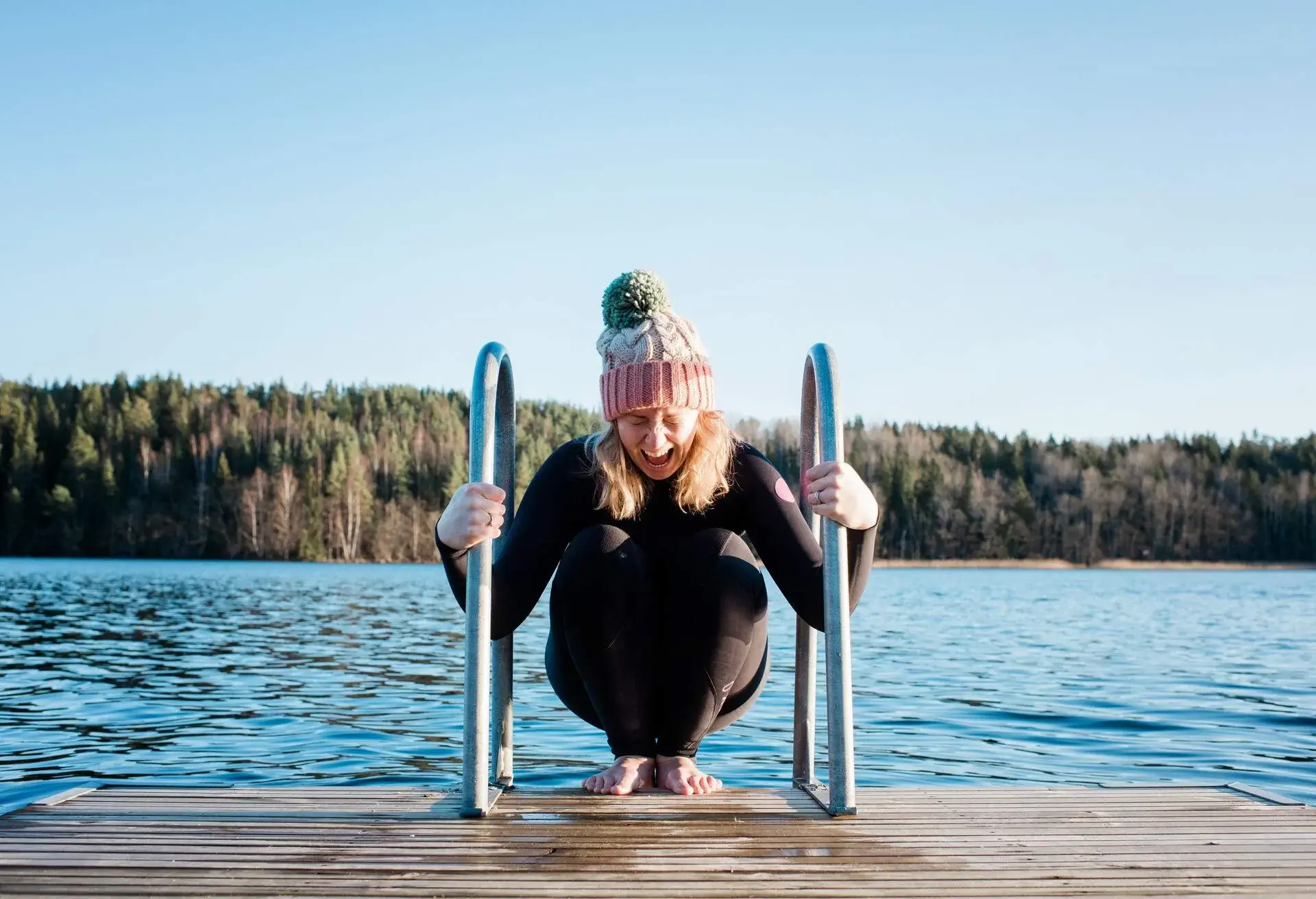 Blonde woman wearing a wool hat and wetsuit about to dip in cold lake in winter