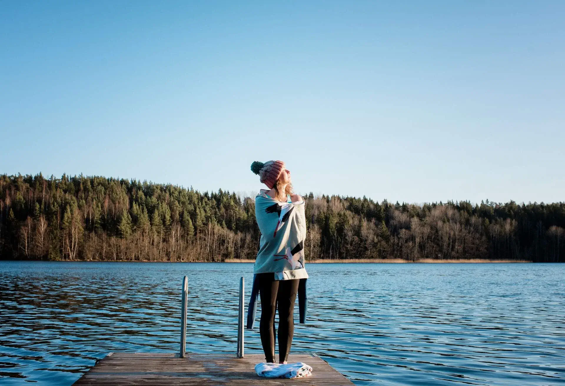 Woman wearing a wool hat, wetsuit and blanket enjoying sun rays by a lake after winter bathing