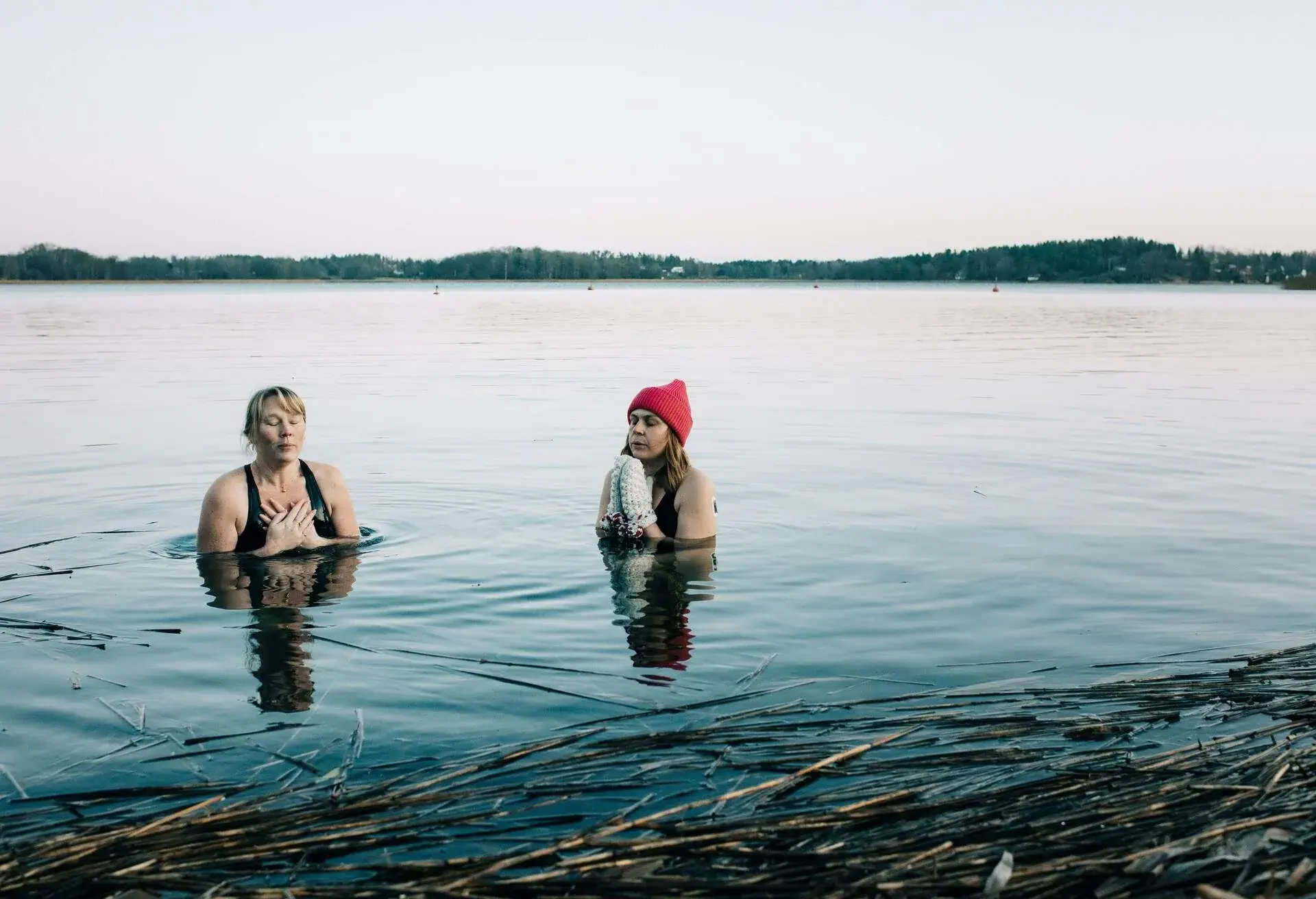 Two women, one wearing wool hat and gloves, dipping into cold water of a lake in winter