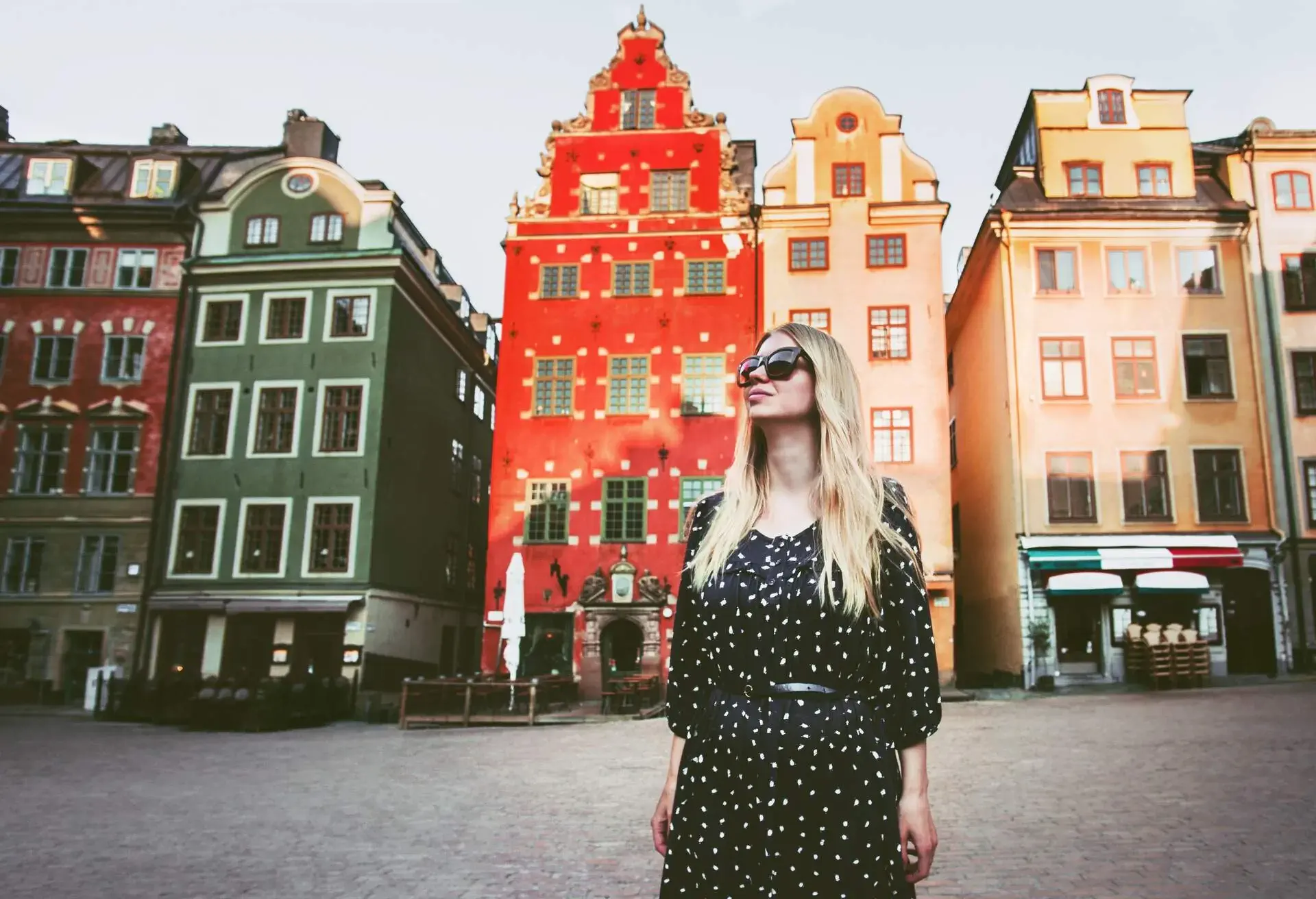 Portrait of blonde woman wearing sunglasses standing in front of a row of colourful tradition north European buildings in town square