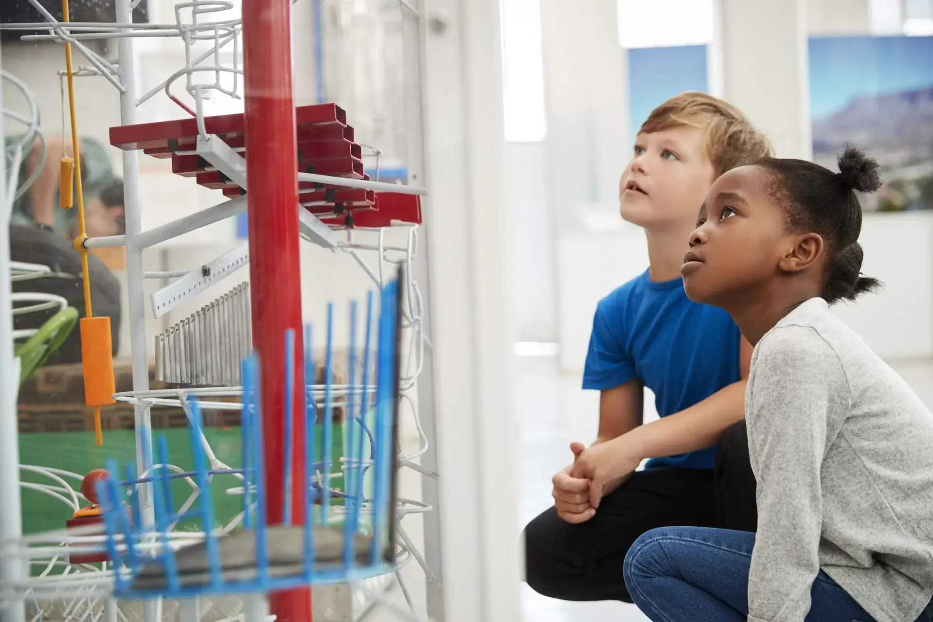 Two kids are examining colourful exhibits on a red pole while squatting.