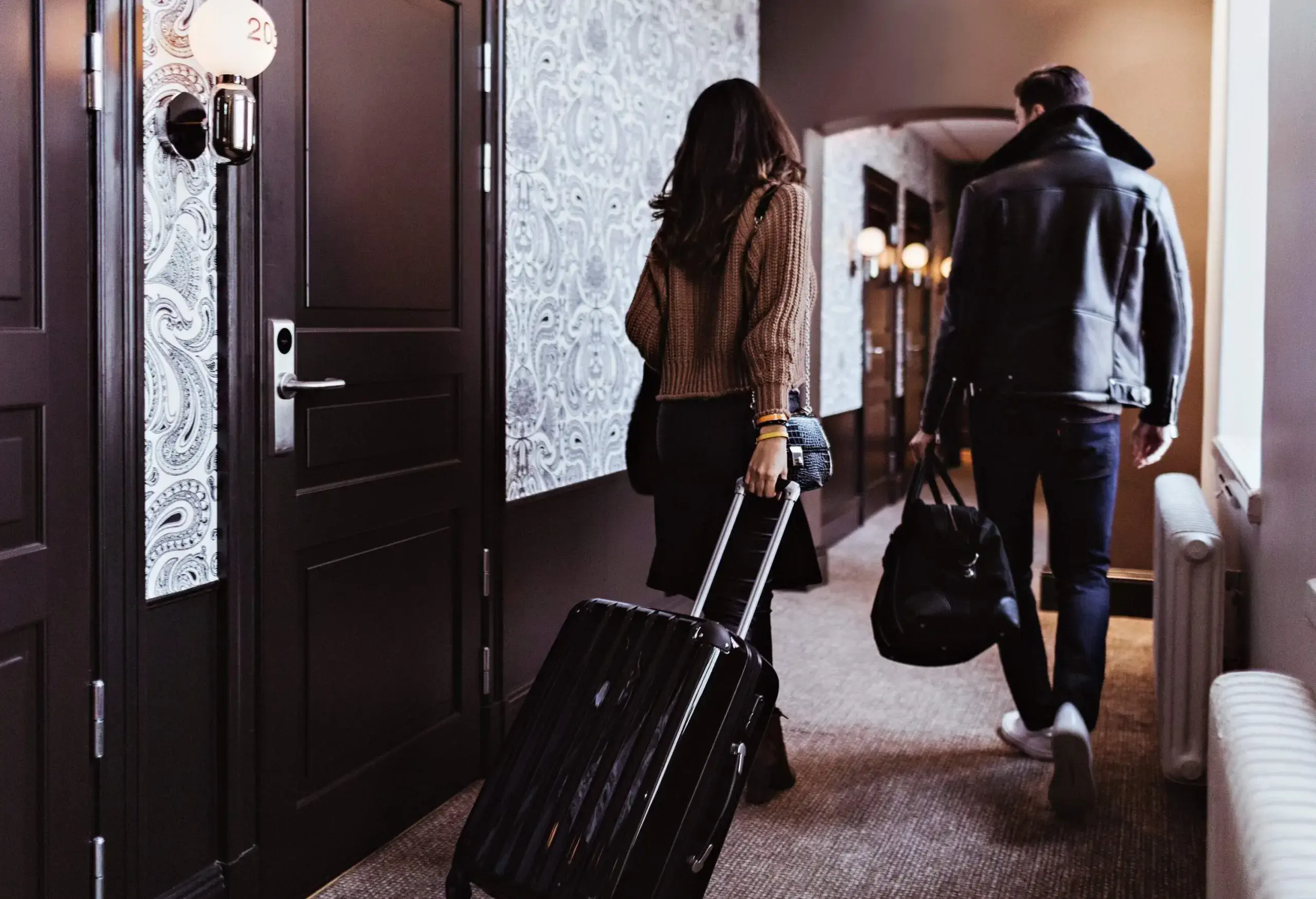 A couple walks down a stylish hotel corridor; one pulls a suitcase while the other carries a bag, both dressed in casual chic attire.