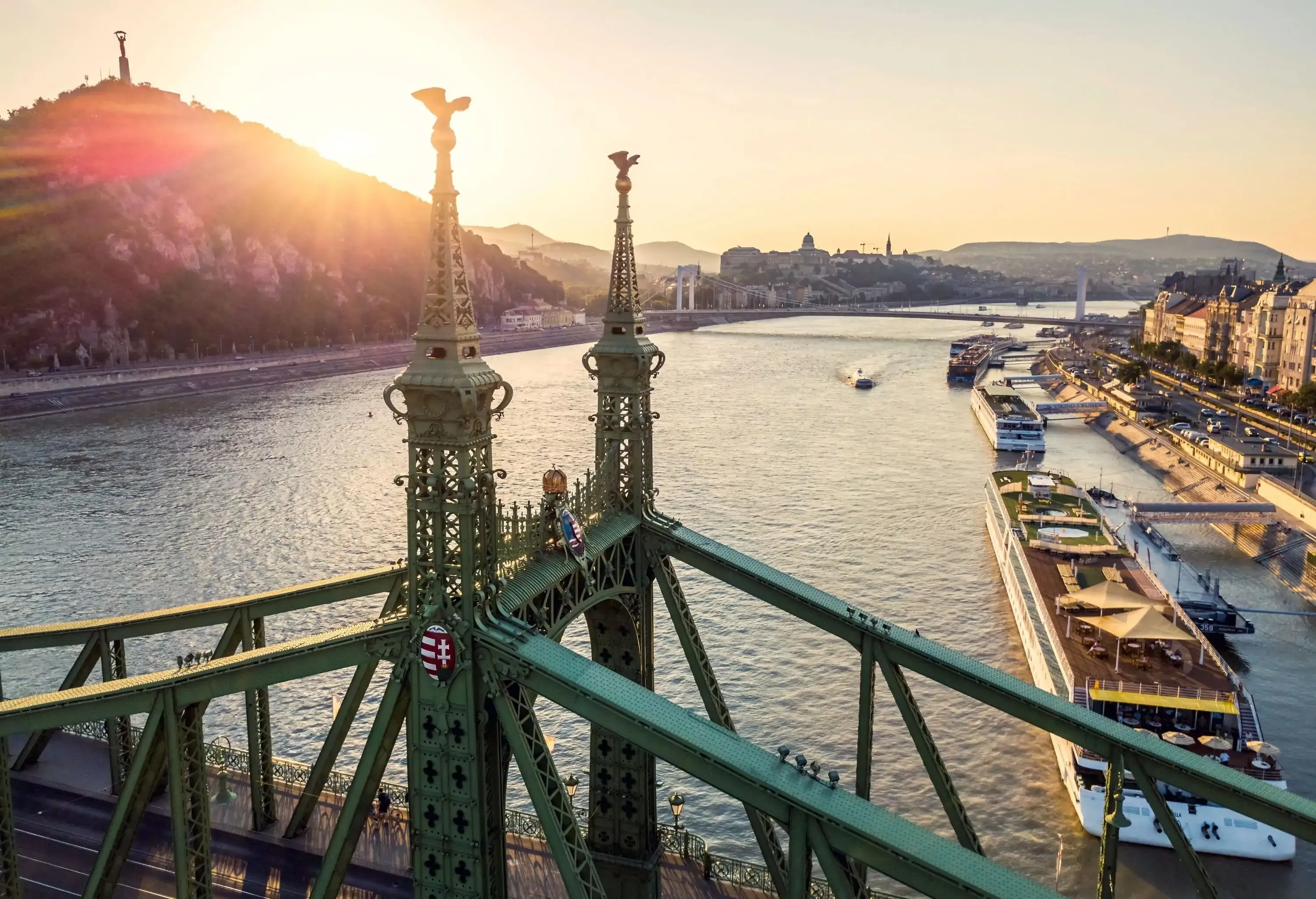 A green, ornate bridge over a wide river at sunset, with boats docked along the riverside and hills silhouetted in the golden light.