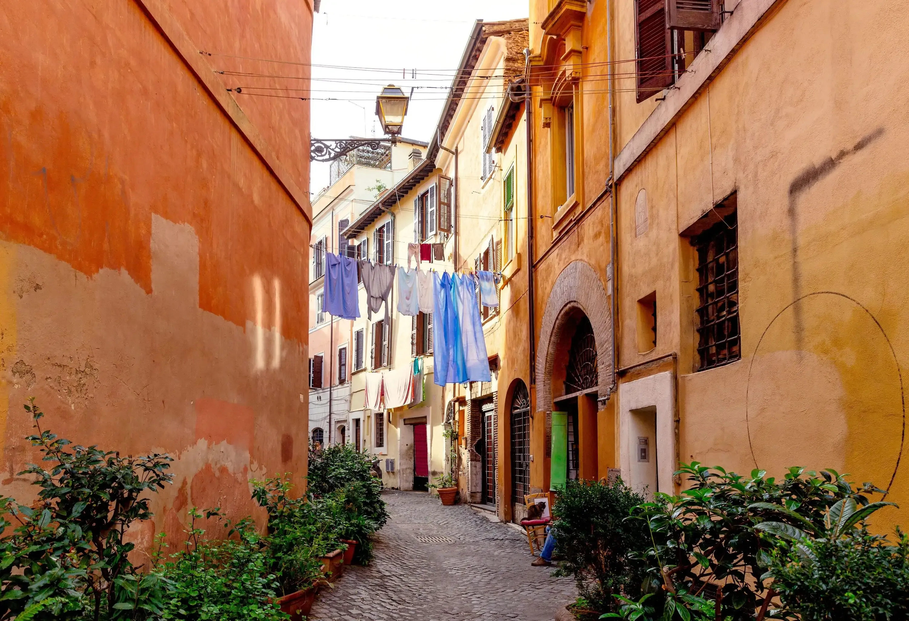 A narrow cobblestone alleyway, flanked by colorful buildings in shades of orange, yellow, and beige. Laundry hangs across the alley on clotheslines, with light blue and white garments swaying gently. Potted plants and greenery line the sides of the path, adding charm to the scene. 