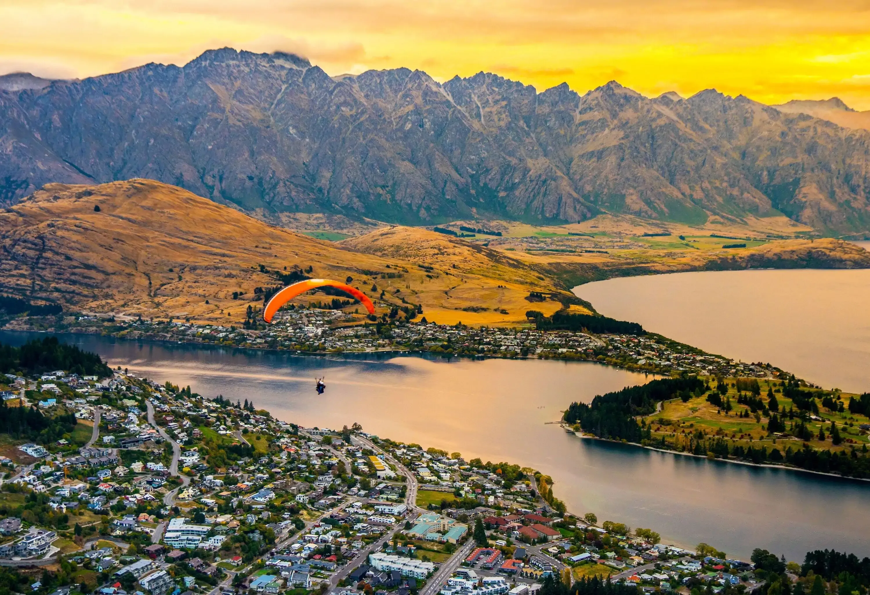 Aerial view of Queenstown, New Zealand, with a paraglider in the sky, Lake Wakatipu, and surrounding mountains under a cloudy sunset.