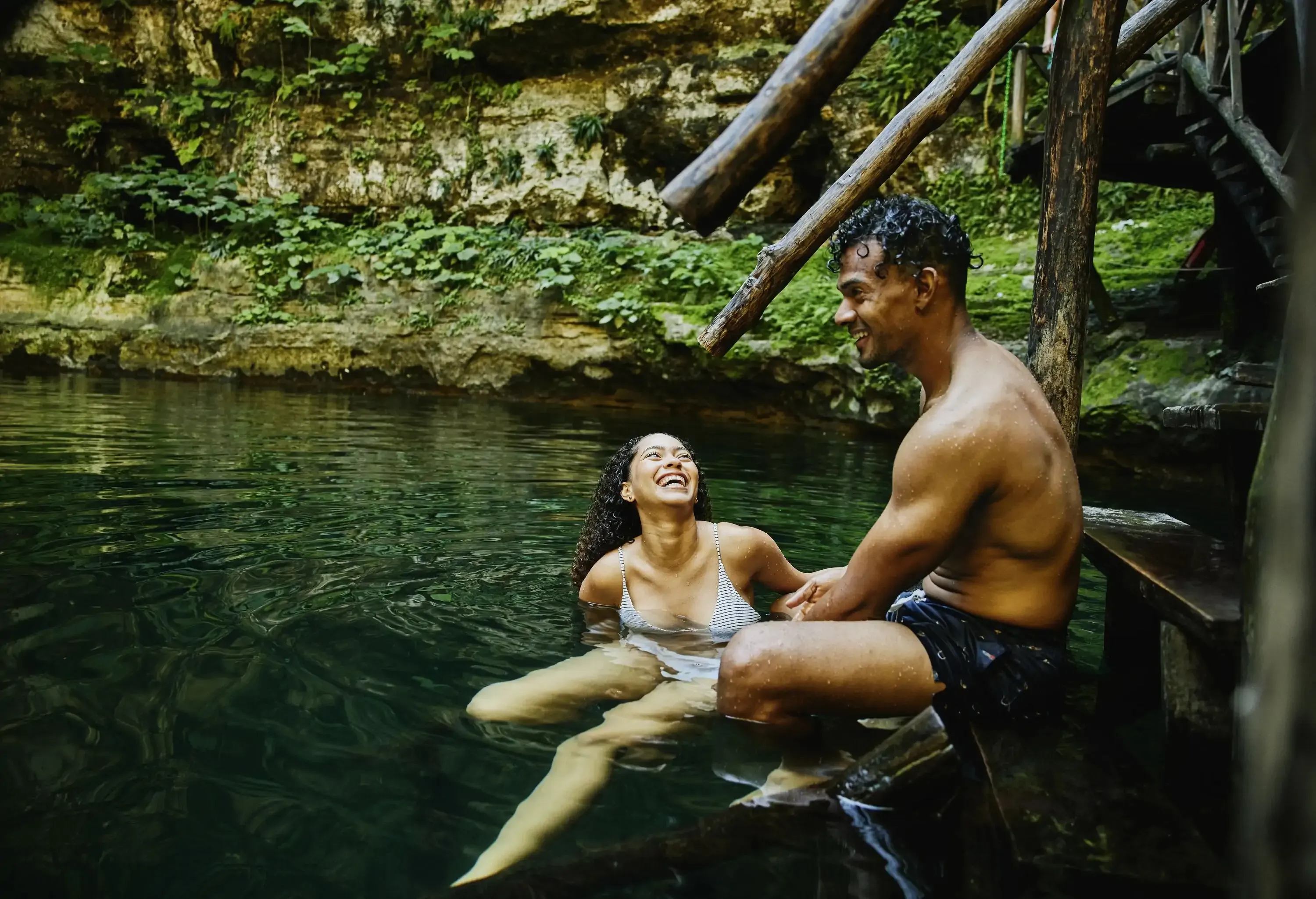 A man and woman enjoy a moment together in a natural setting, sitting by the edge of a serene green water body surrounded by lush foliage and rocky terrain. The woman is laughing, leaning slightly back, while the man smiles warmly at her. Wooden structures and steps lead into the water.