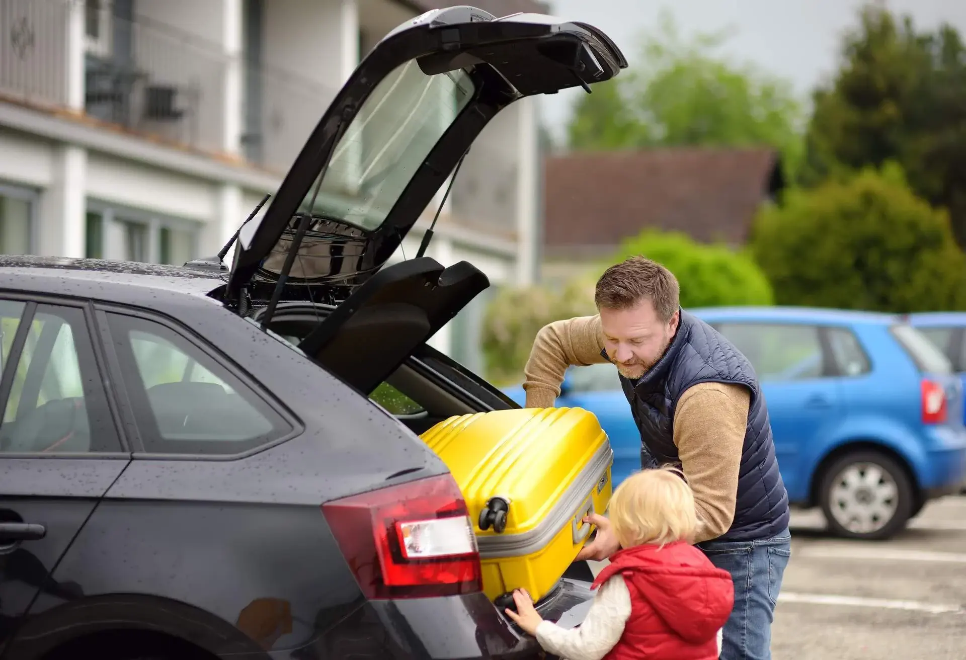 A man putting a suitcase in the trunk of a car, with a child helping him