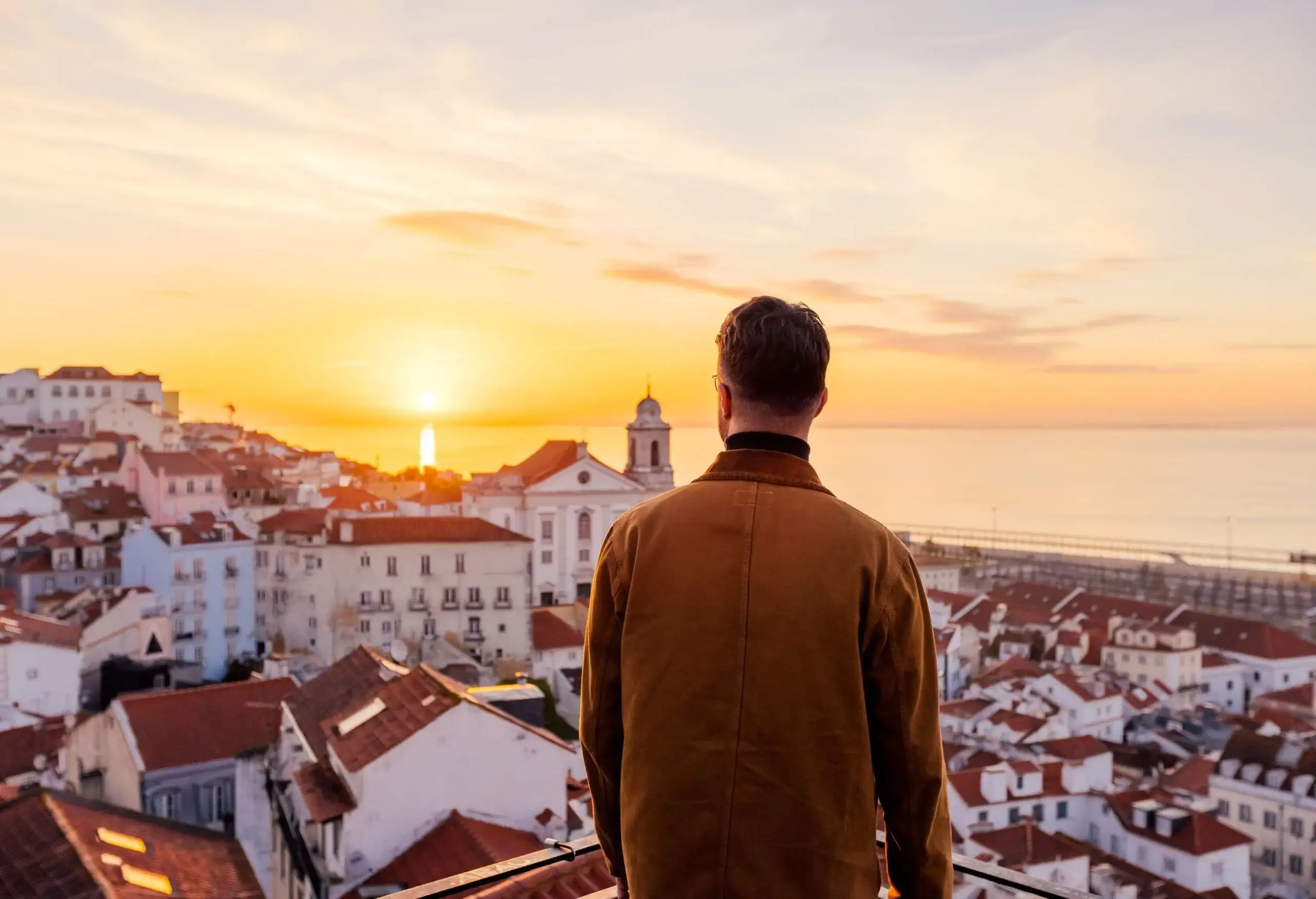 A person's back view, wearing a jacket, gazing out over a picturesque town adorned with charming white houses topped with brick roofs.