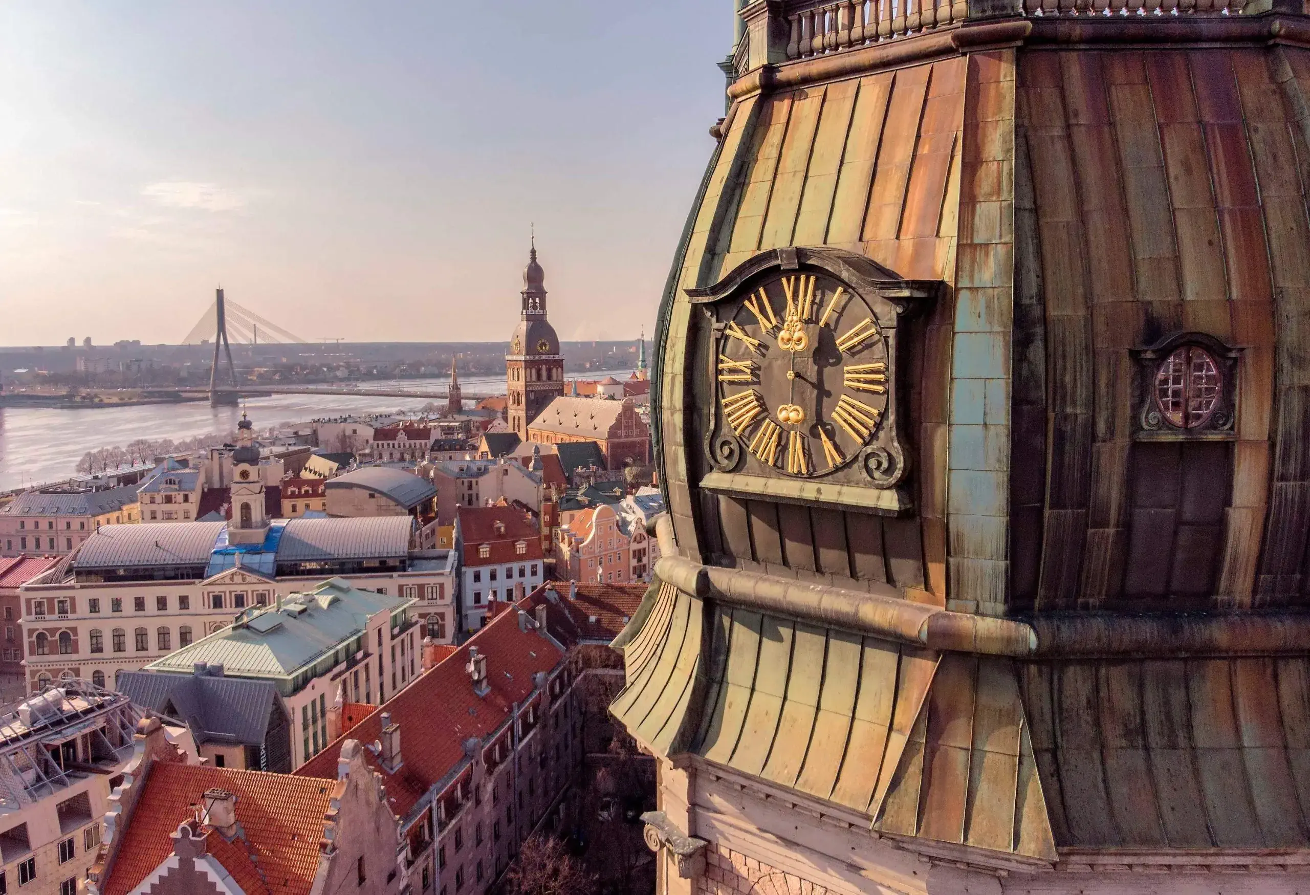 A close-up view of a clock on a church tower overlooking the river along the compact buildings and spanned by a cabled stayed bridge.