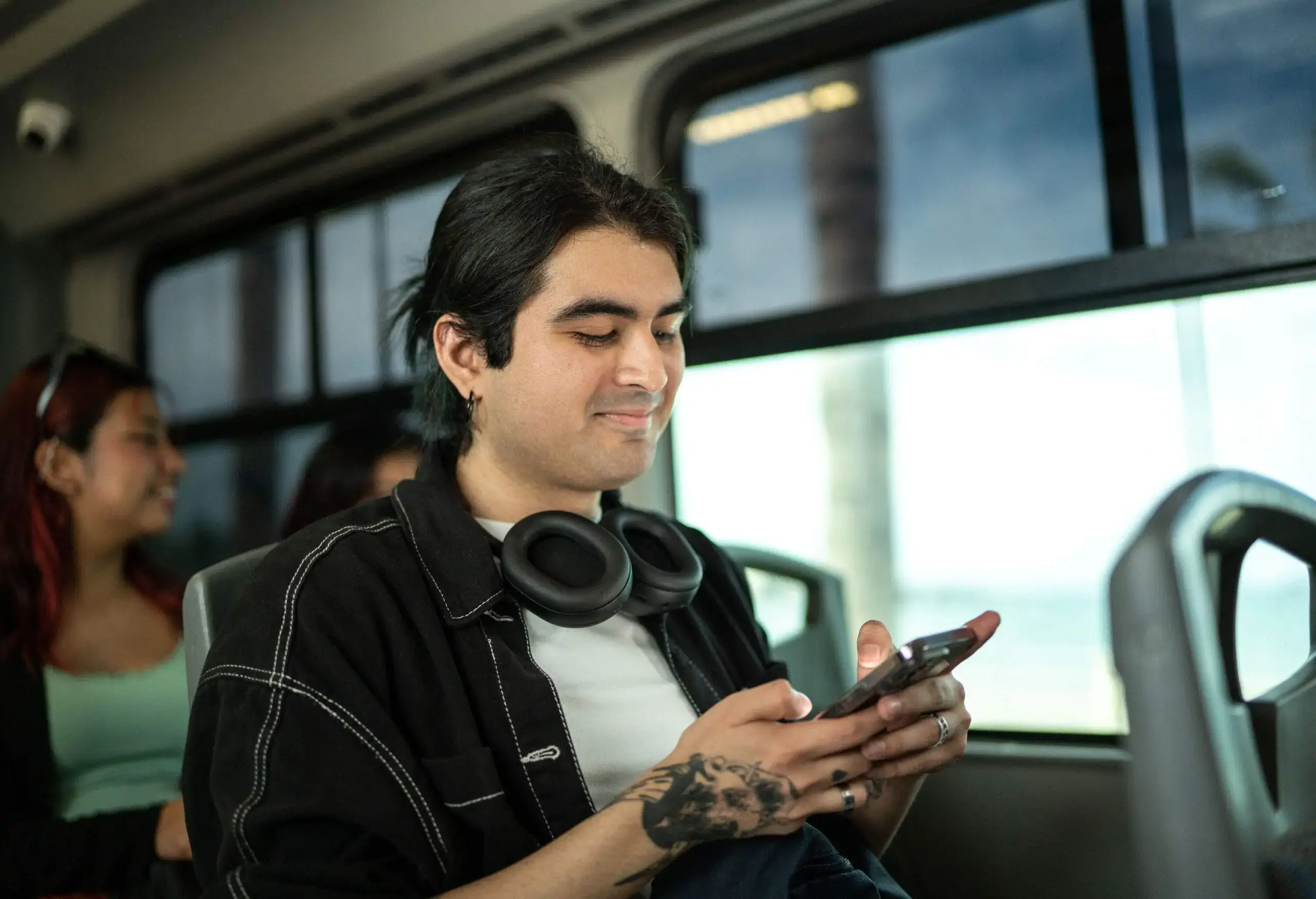 A young man on a bus wearing headphones around his neck and totally absorbed in his smartphone.