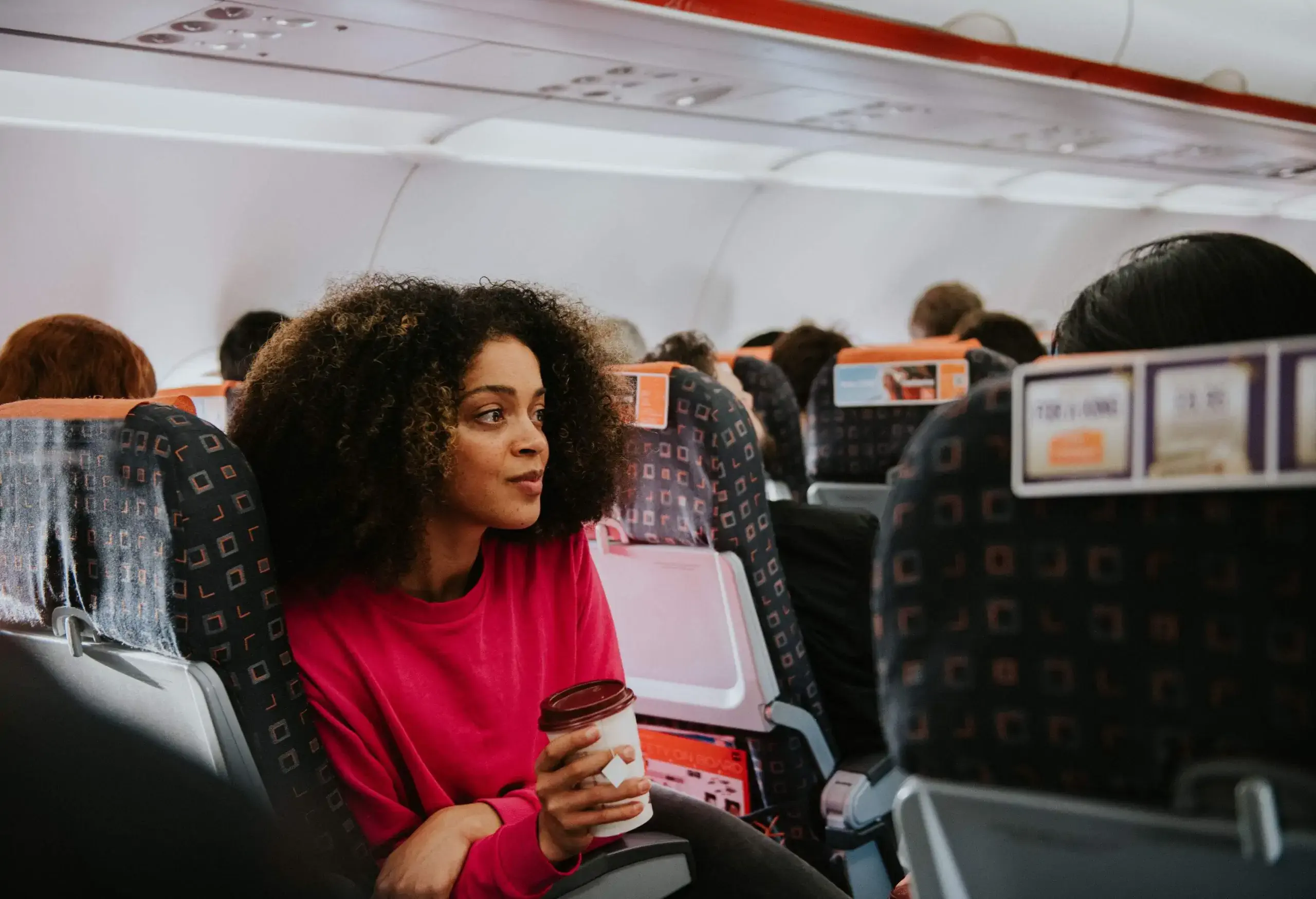 A young woman sits on a crowded plane on an aisle seat. She clutches a cup of coffee and looks perplexed and thoughtful.