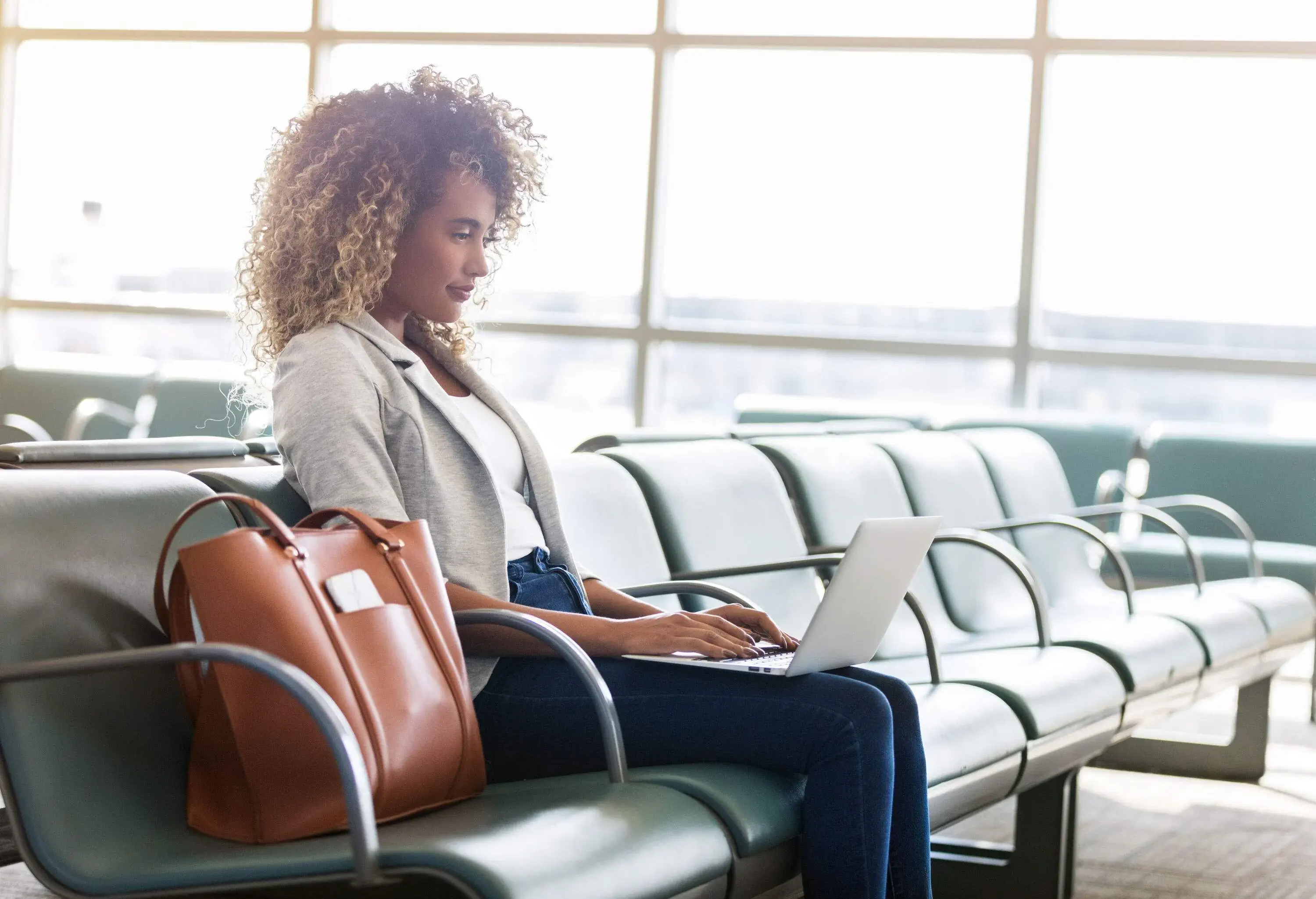 A woman working on her laptop at the airport, her brown bag on a chair next to her.