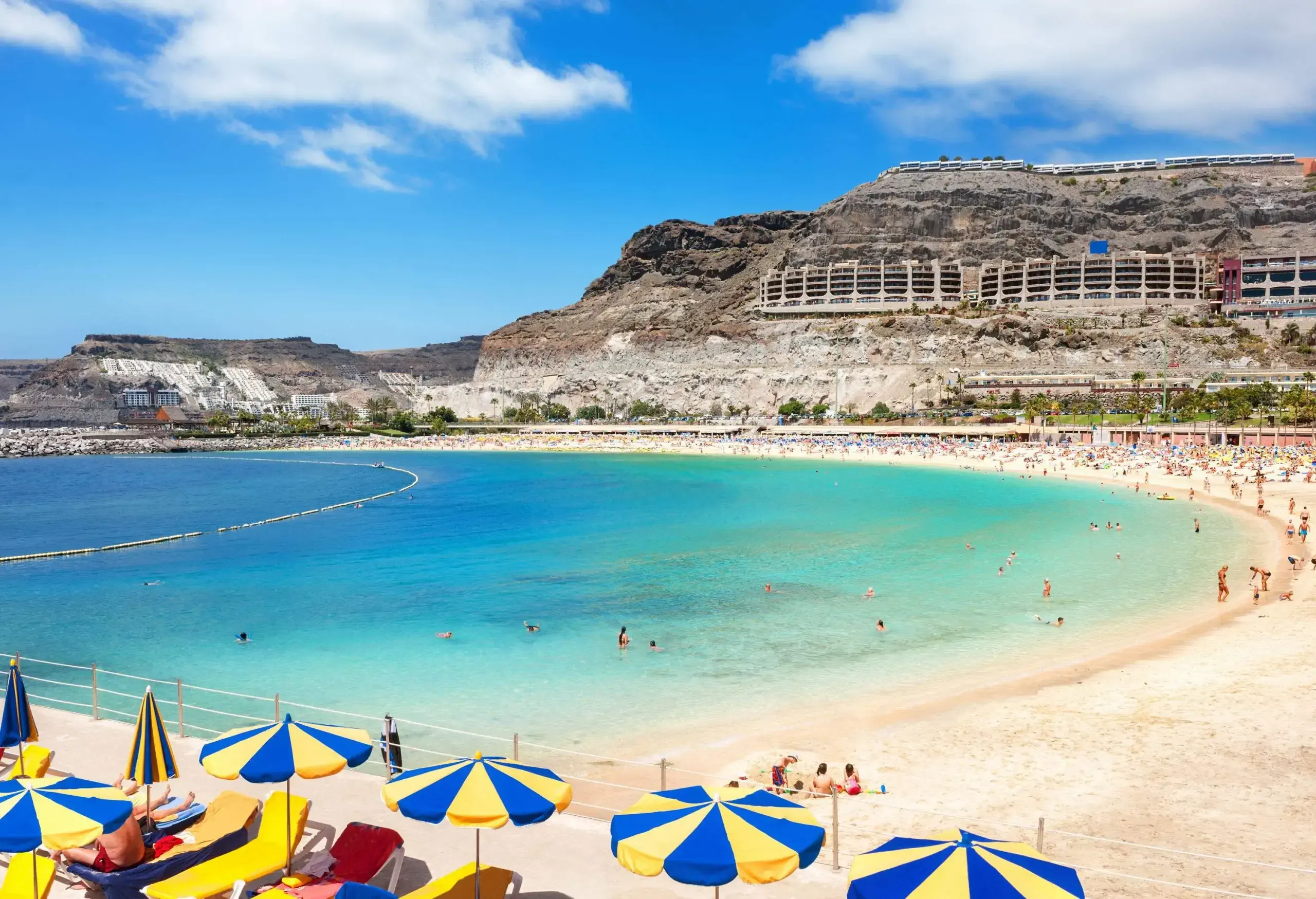 A crowd of people and colourful parasols on the shore of the tranquil clear water beach.
