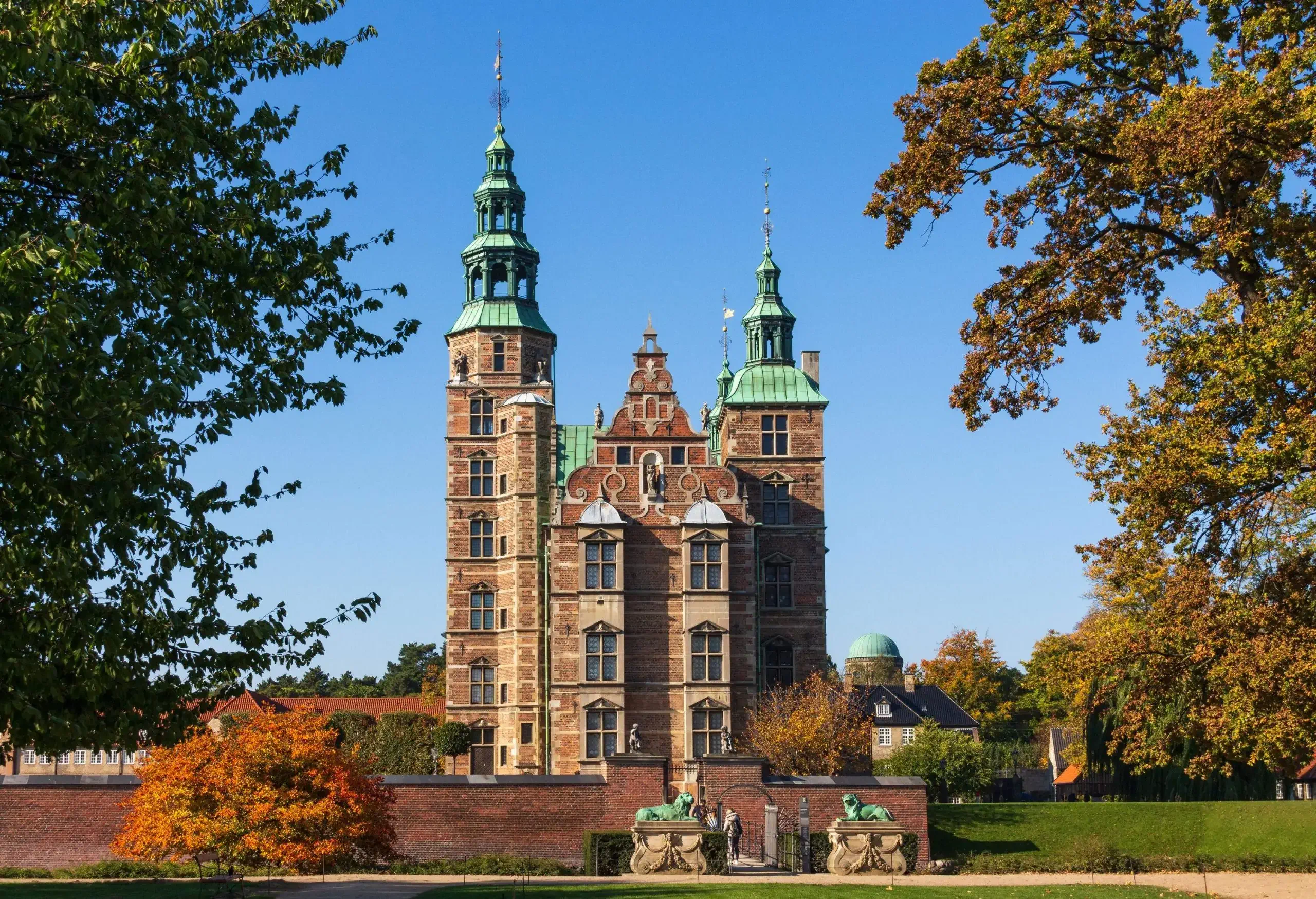 Rosenborg Castle is a brown Renaissance-style castle surrounded by a concrete gate and two towering spires of varying heights.