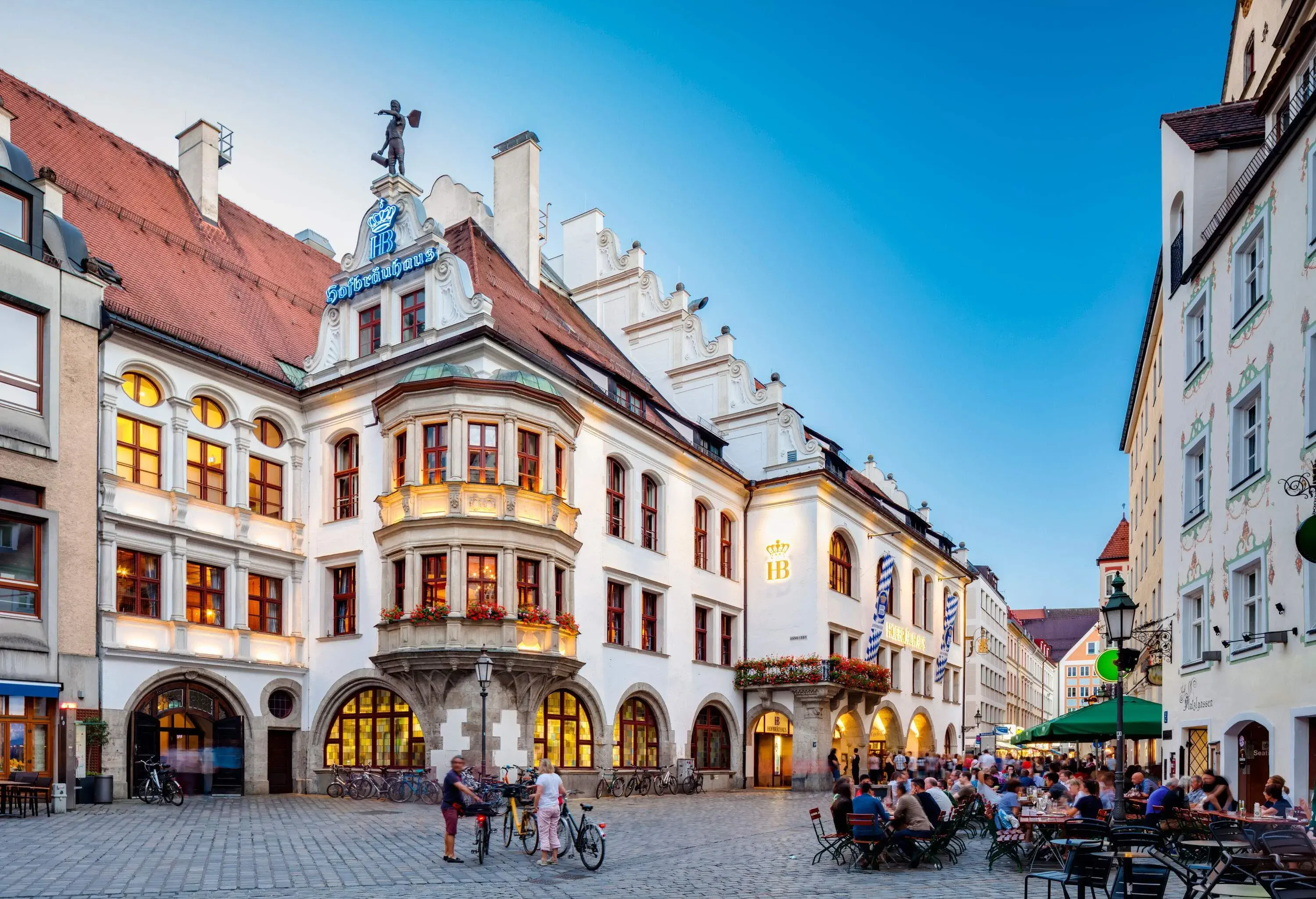 A row of buildings, one of which has "Hofbrauhaus" spelled out on the front roof edge of the building facing an outdoor cafe.
