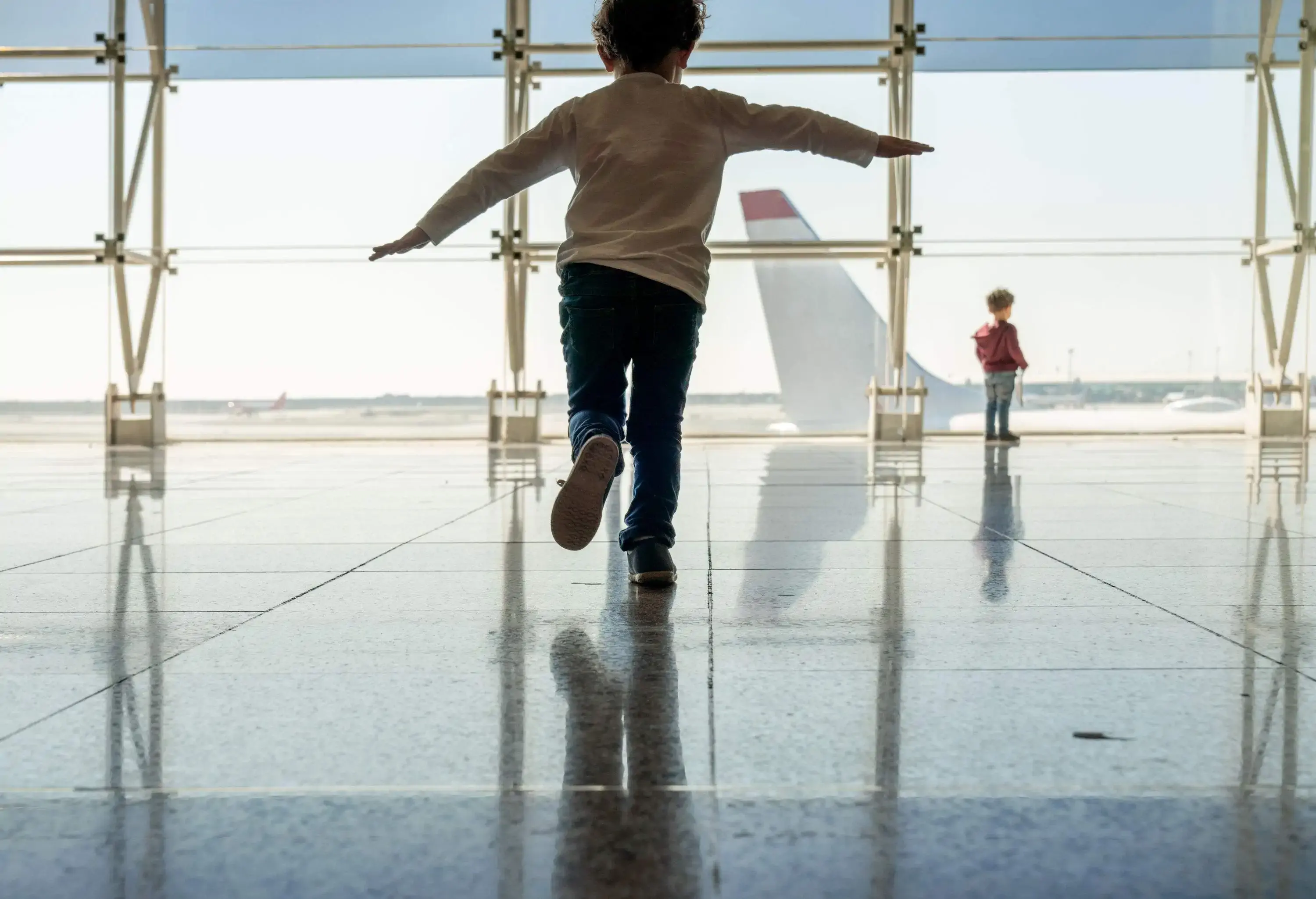 A boy balancing on one foot and another on the glass window looking at the airplane in the airport.