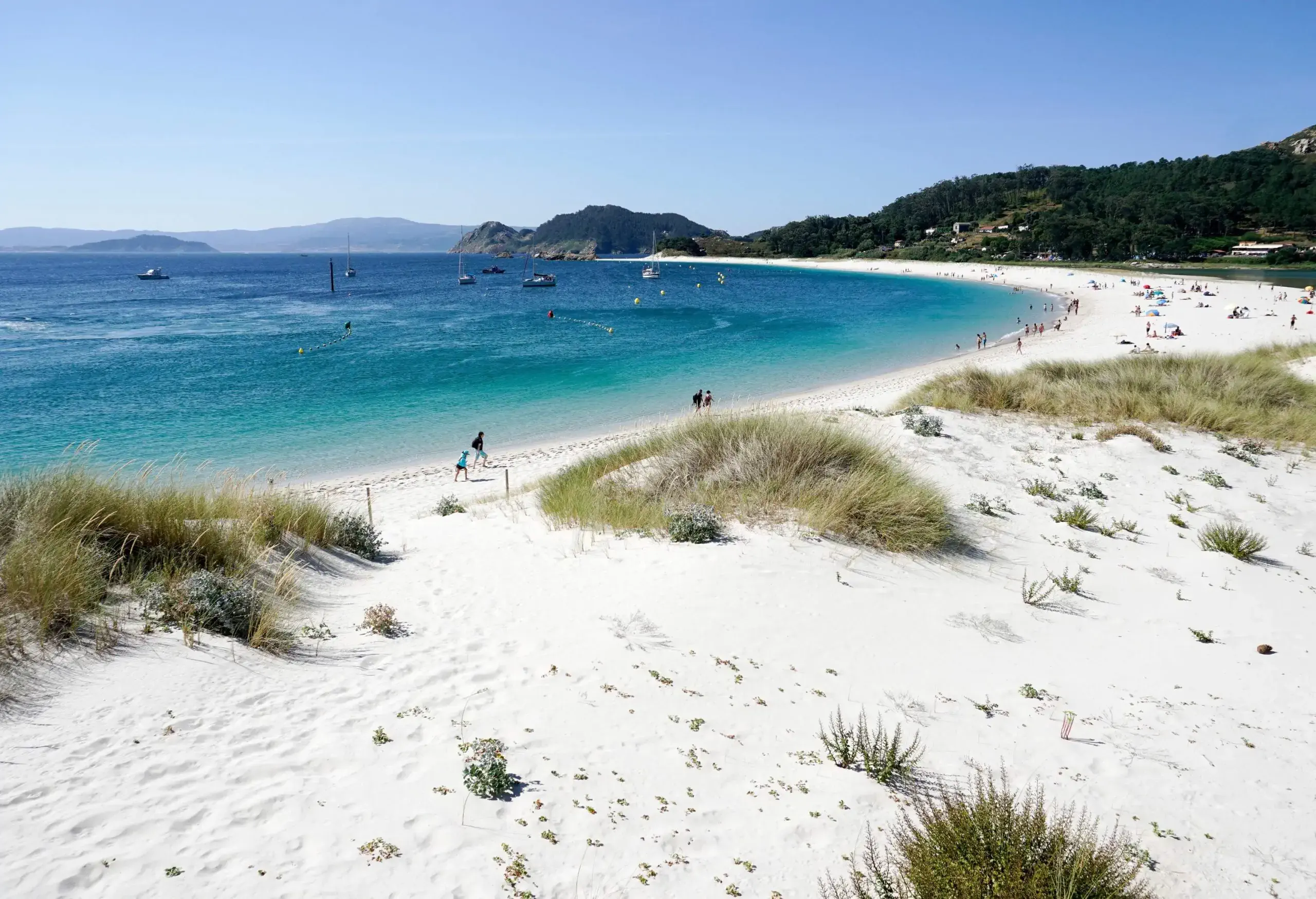 A white sand beach with boats and people is seen at the base of a lush, forested hill.