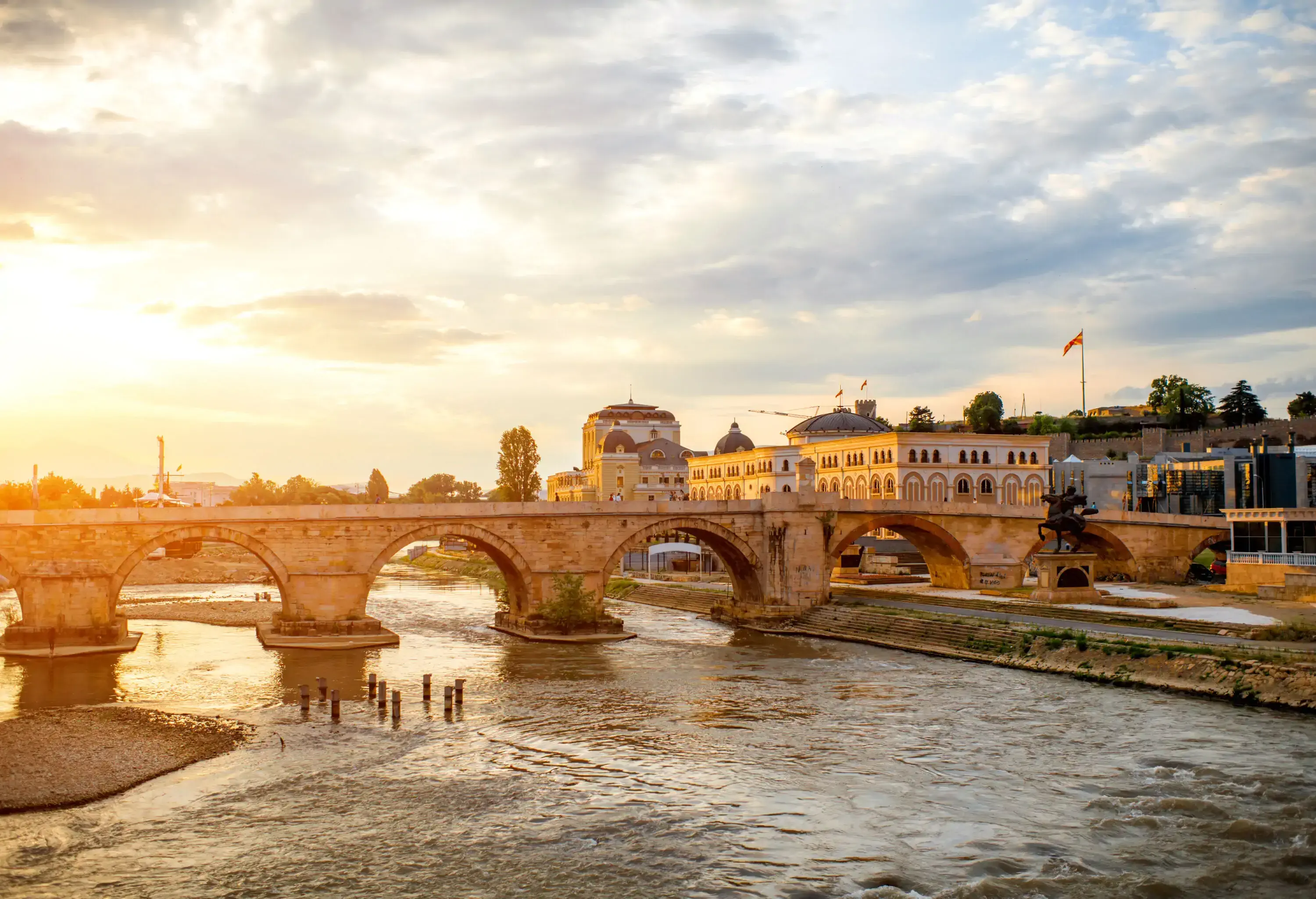 The scenic sun shines through a stone arch bridge that spans the river along the old buildings.