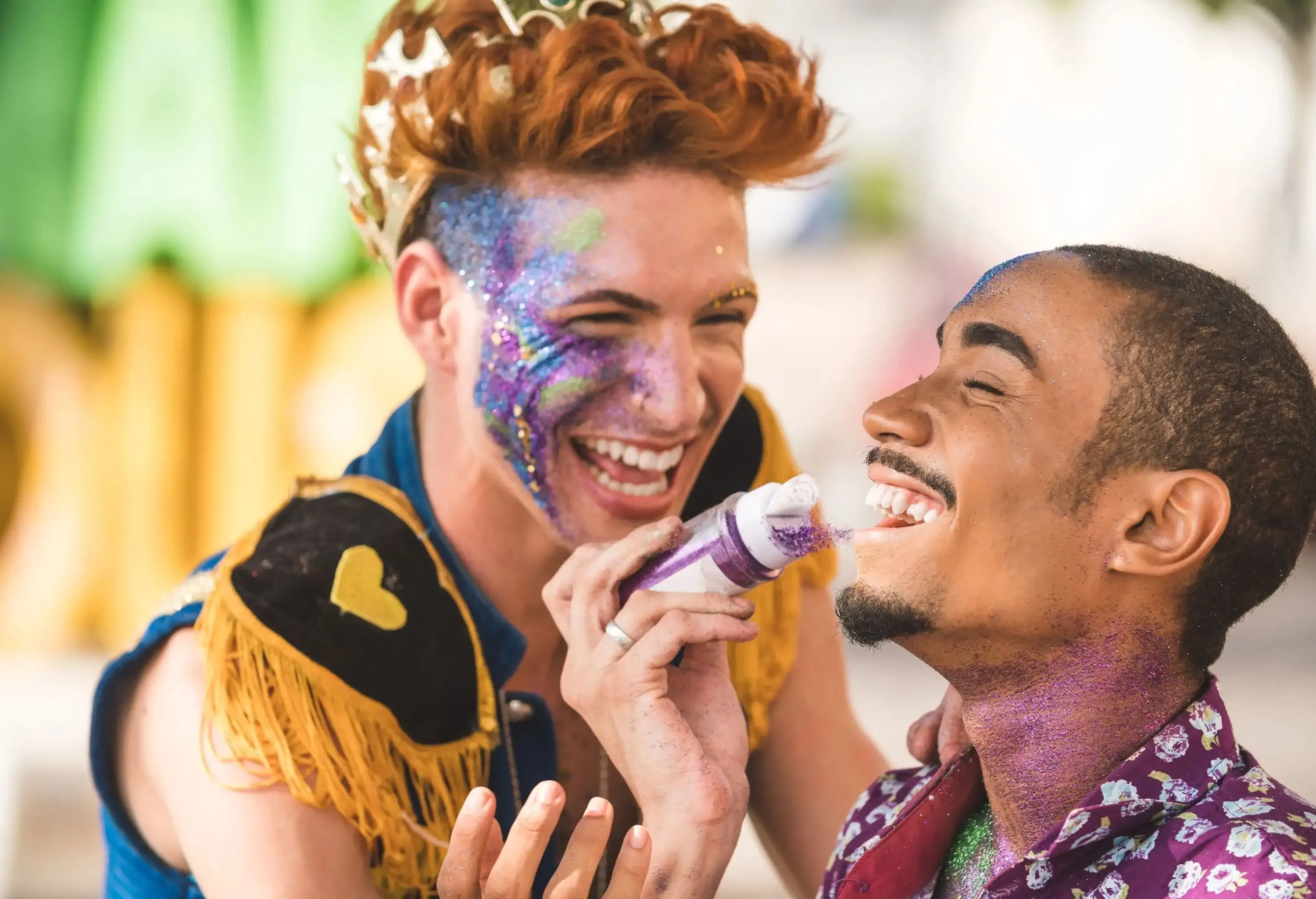 A man with orange hair, a colourful costume, and glossy makeup laughs as he applies glitter to a man's face.