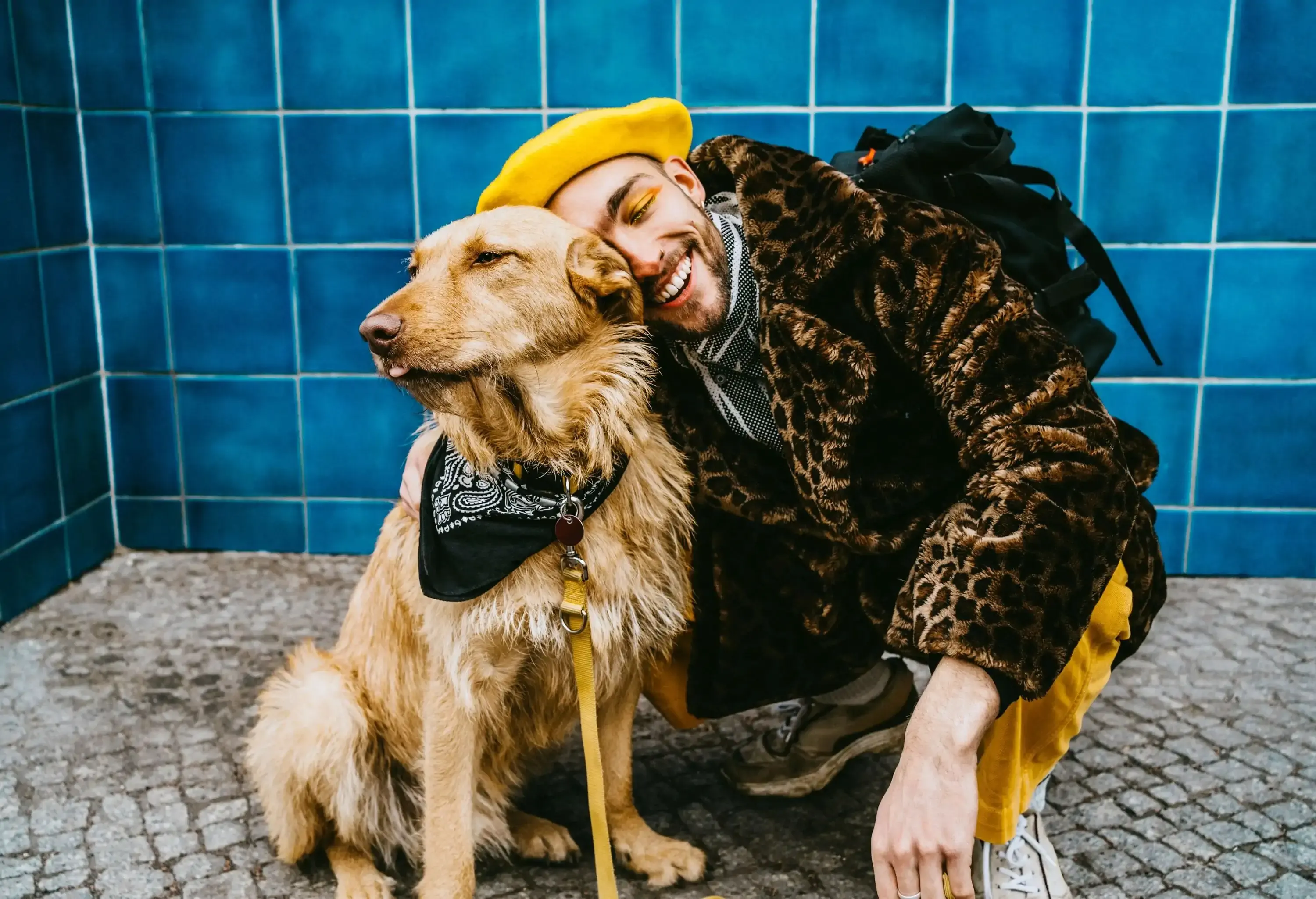 A man in an animal print jacket and yellow beret smiles as he hugs a brown dog with a black scarf.