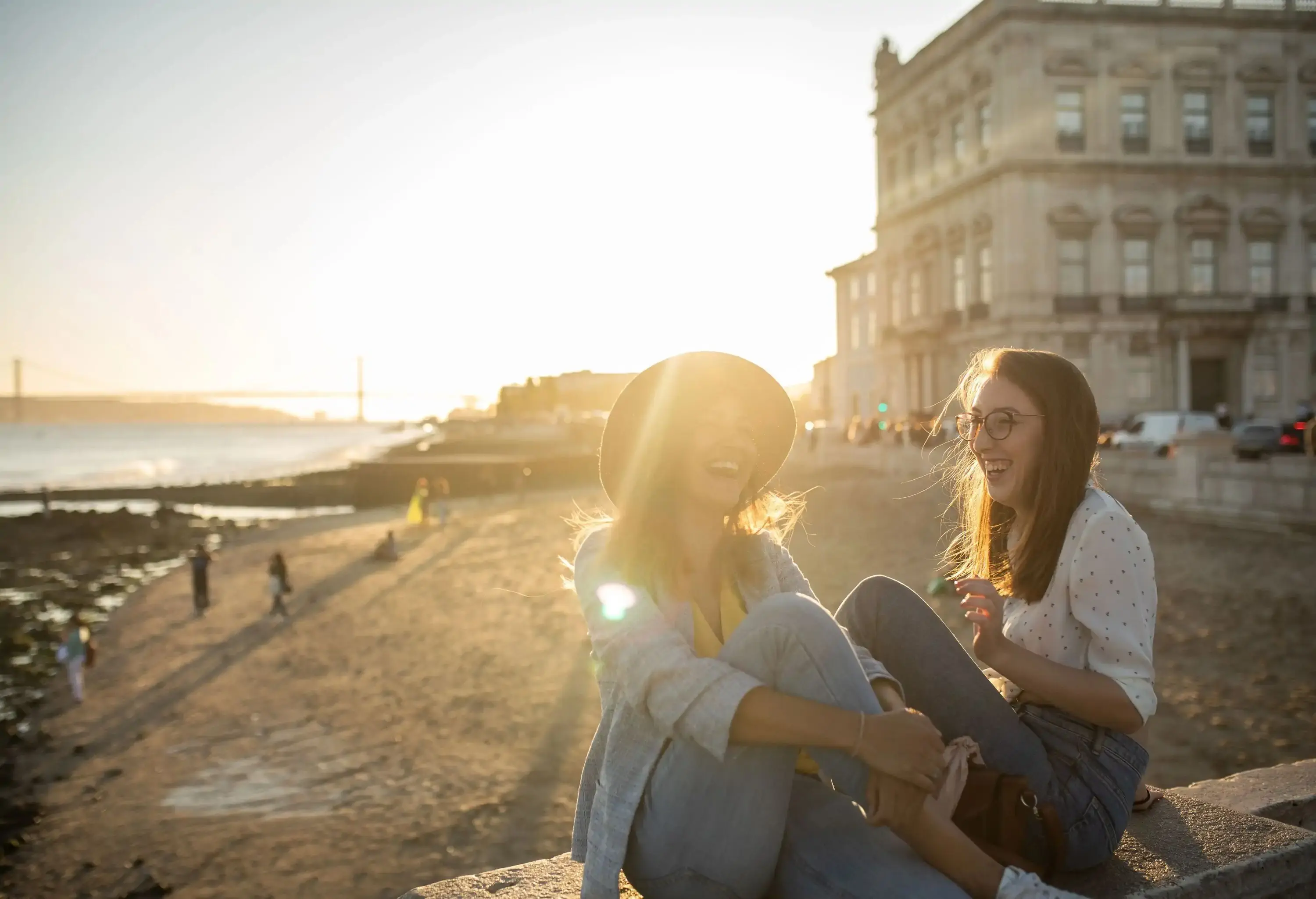 Two laughing women during a conversation while seated on a bridge over the shore on a sunny day outdoors.