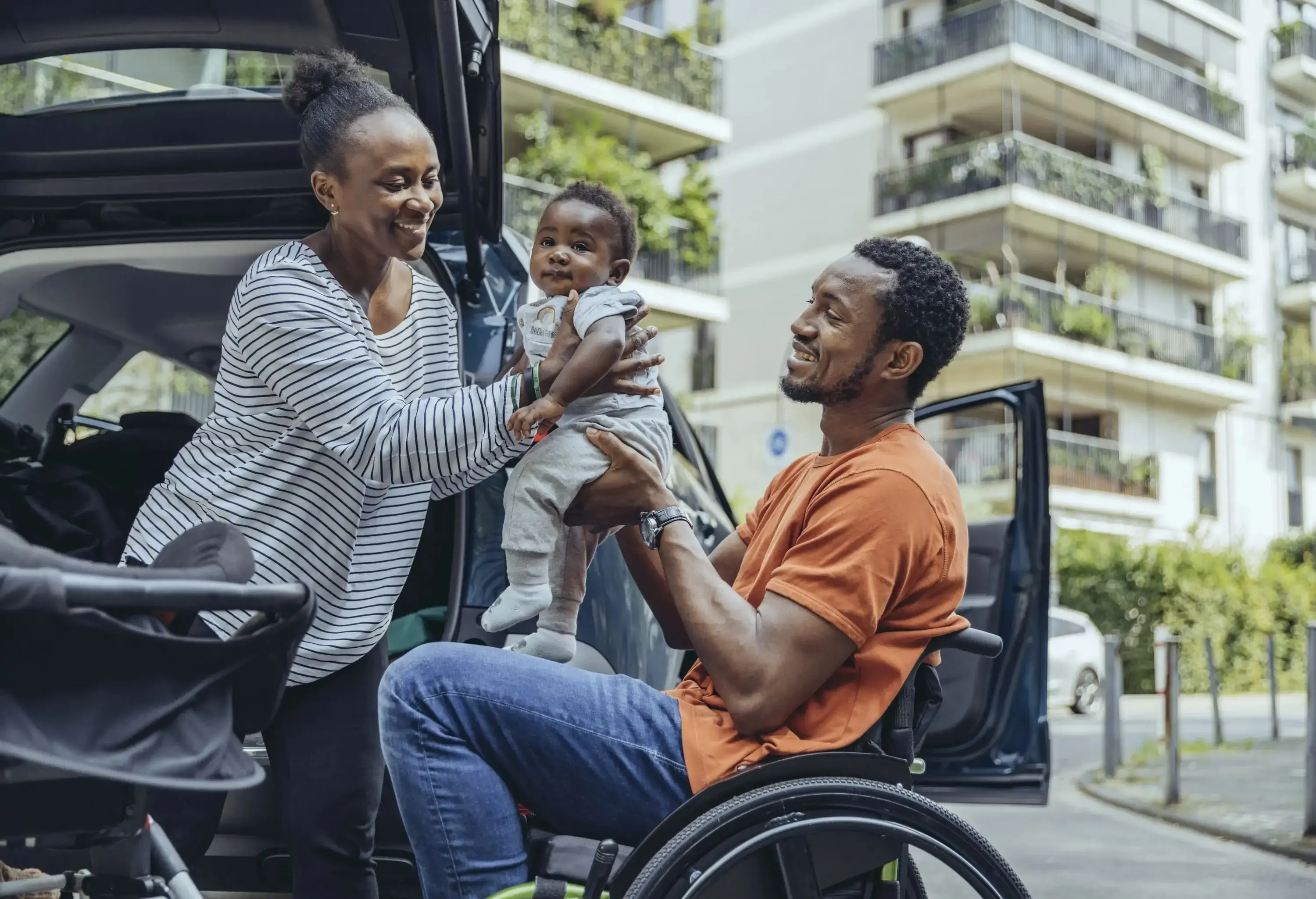 Mother giving son to father sitting in wheelchair by car