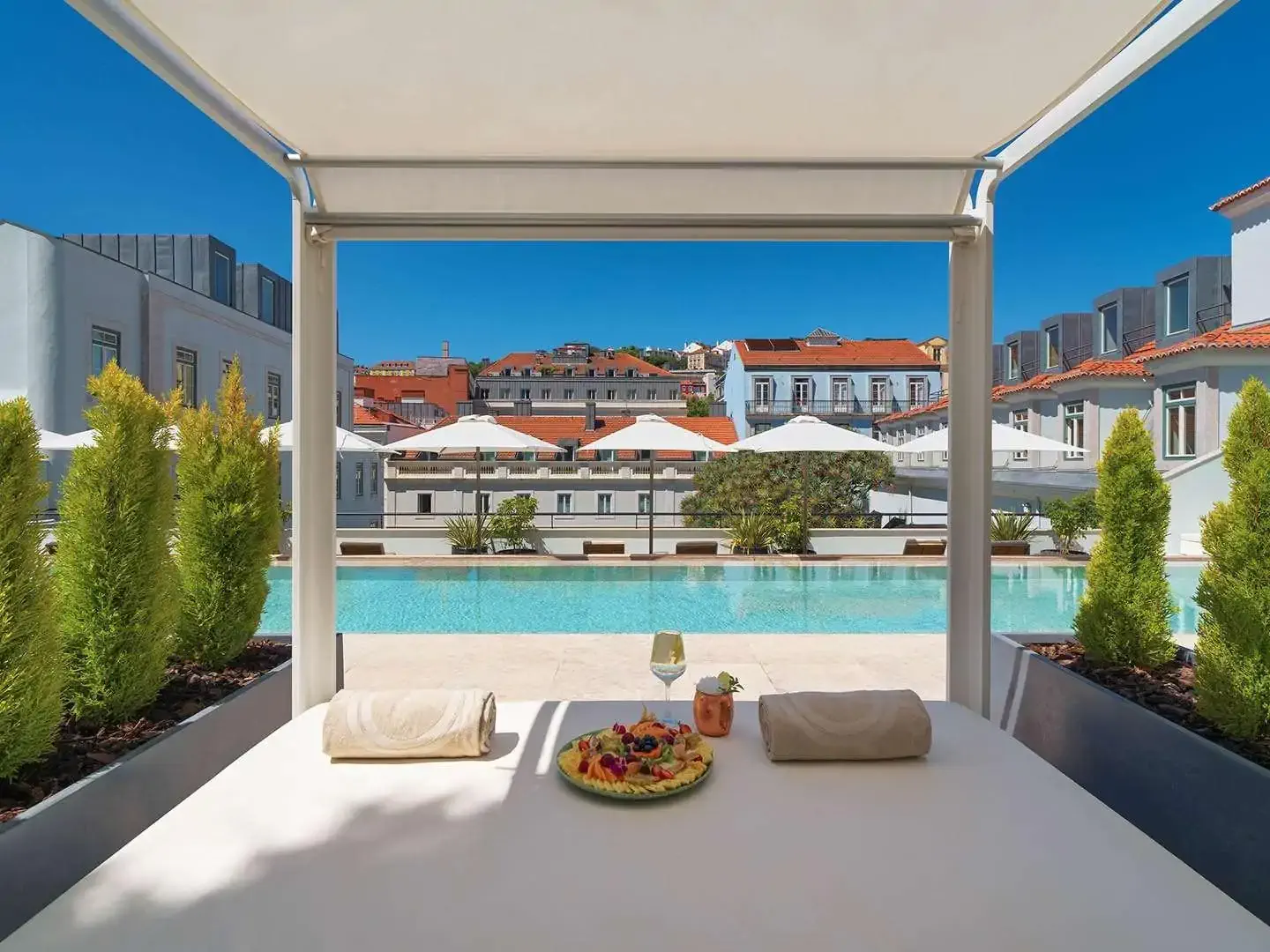 A serene poolside cabana with a shaded daybed, a refreshing drink, and a fruit platter overlooking a sparkling blue pool.