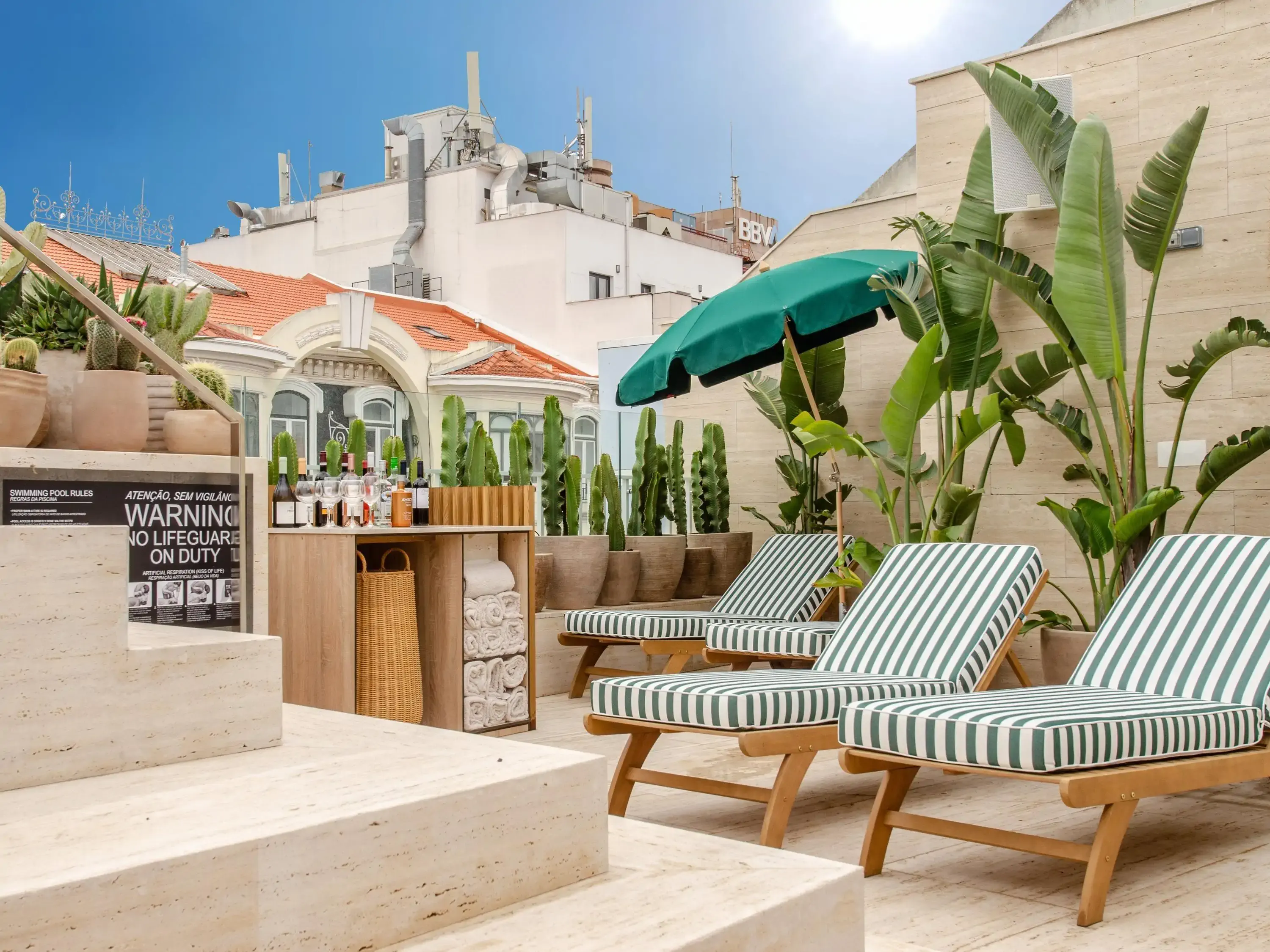 A stylish rooftop terrace with striped lounge chairs, tropical plants, and a bar area set against a backdrop of city rooftops.