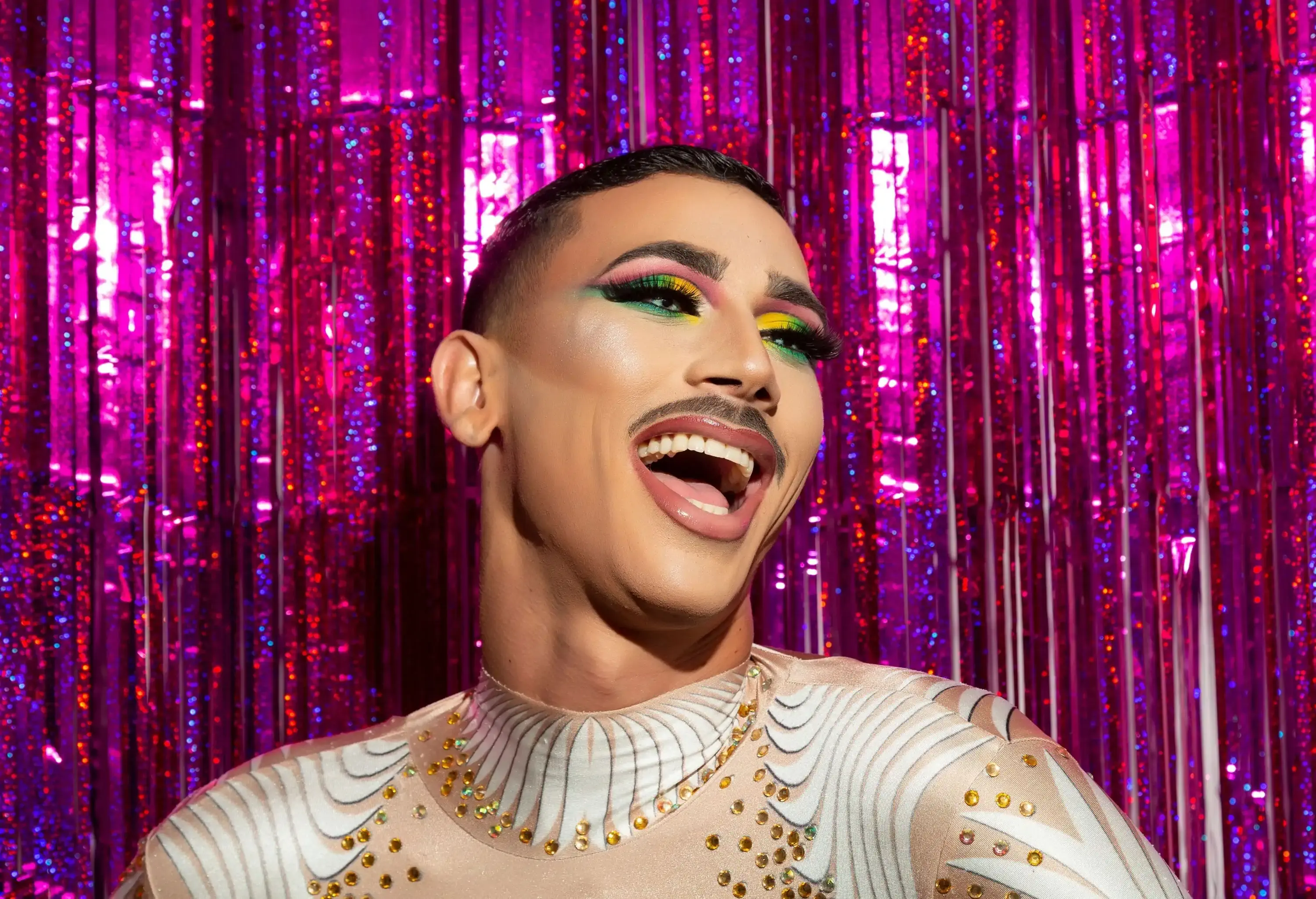 A radiant young gay man wearing makeup beams with joy, mouth open in delight, against a lively and colourful backdrop.