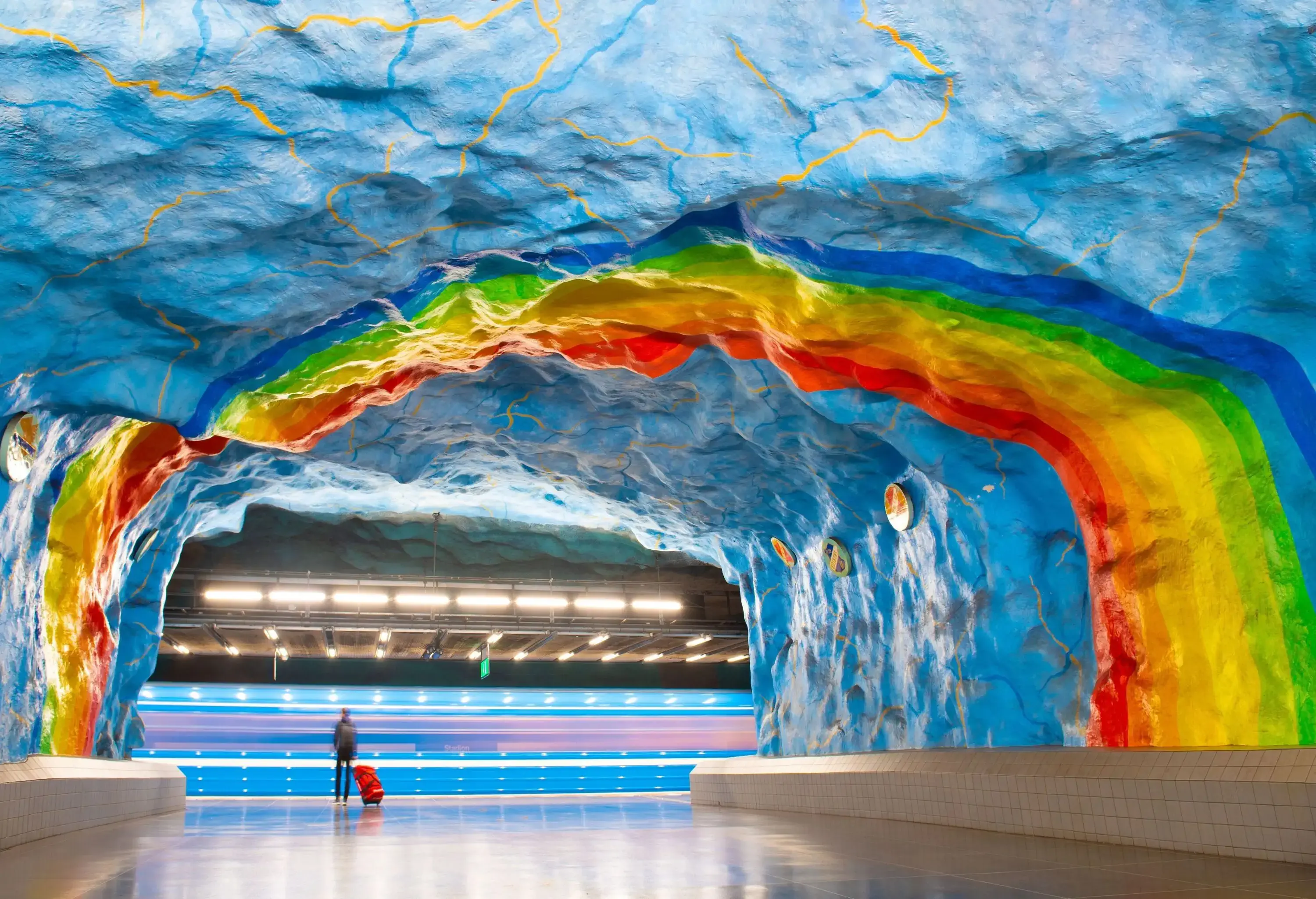 A person with luggage stands on a subway train platform under an arched ceiling of rainbow-coloured jagged rocks.