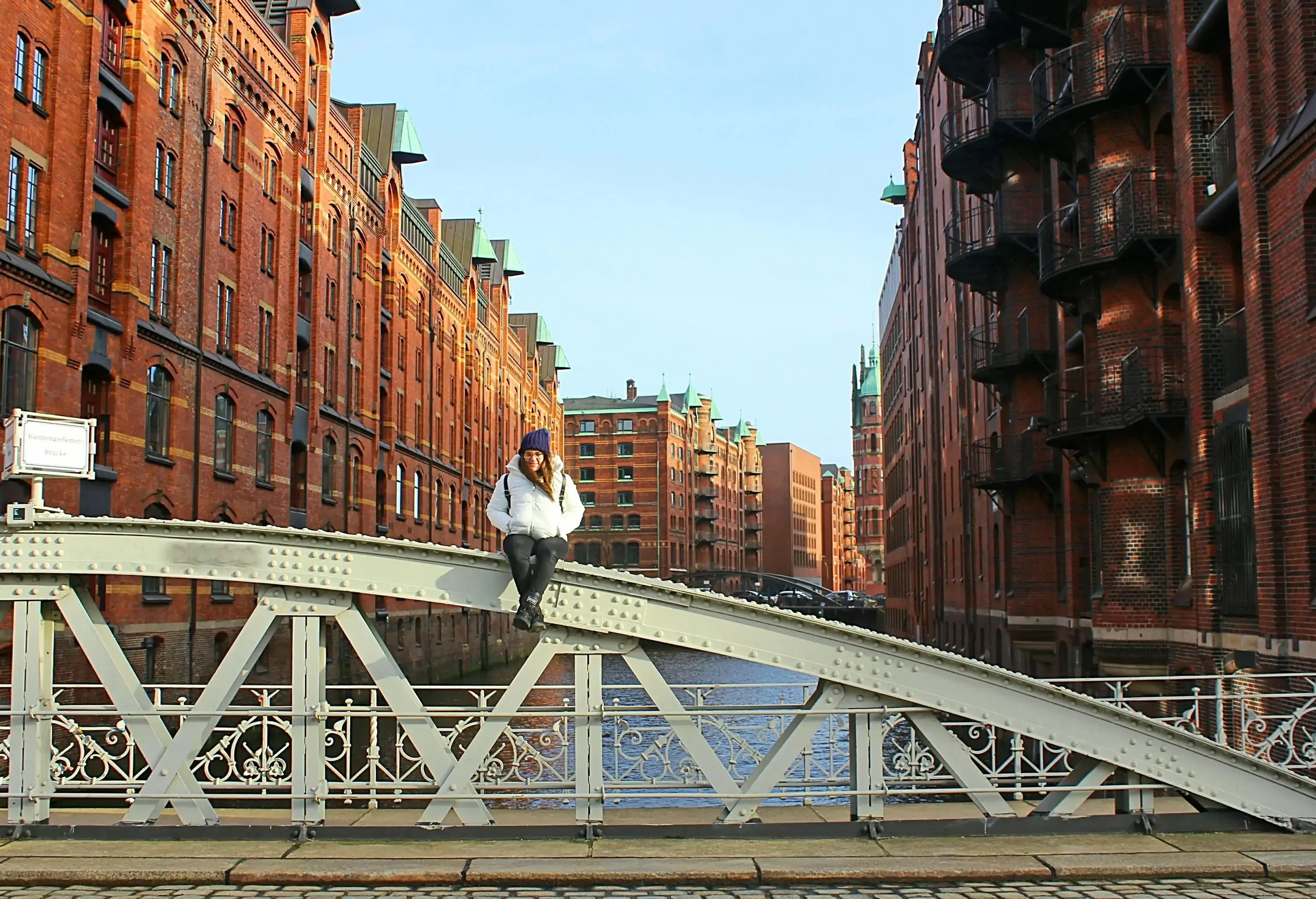 A woman sits on an iron arch fence of a bridge with tall buildings in the background.
