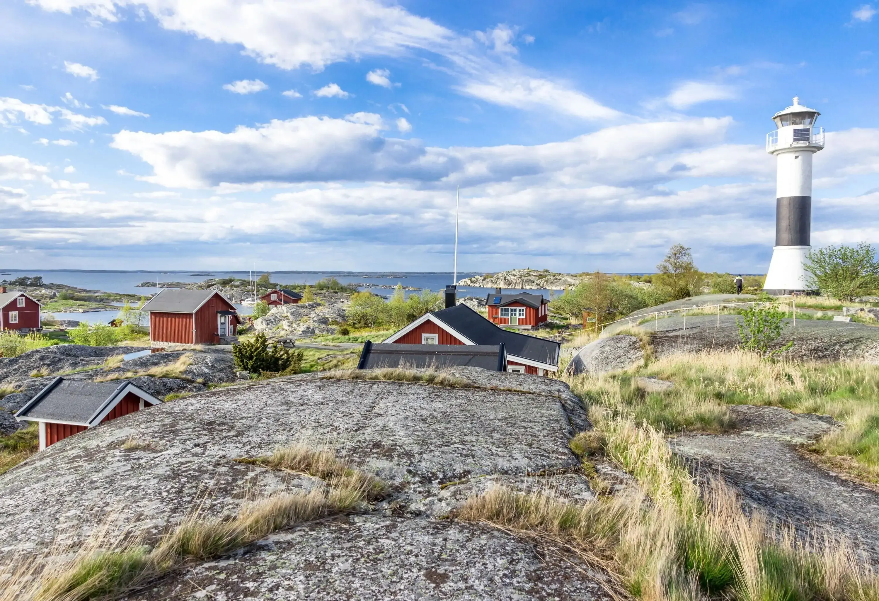 Traditional houses on the rocky shore of a lake with a white lighthouse against the cloudy blue sky.
