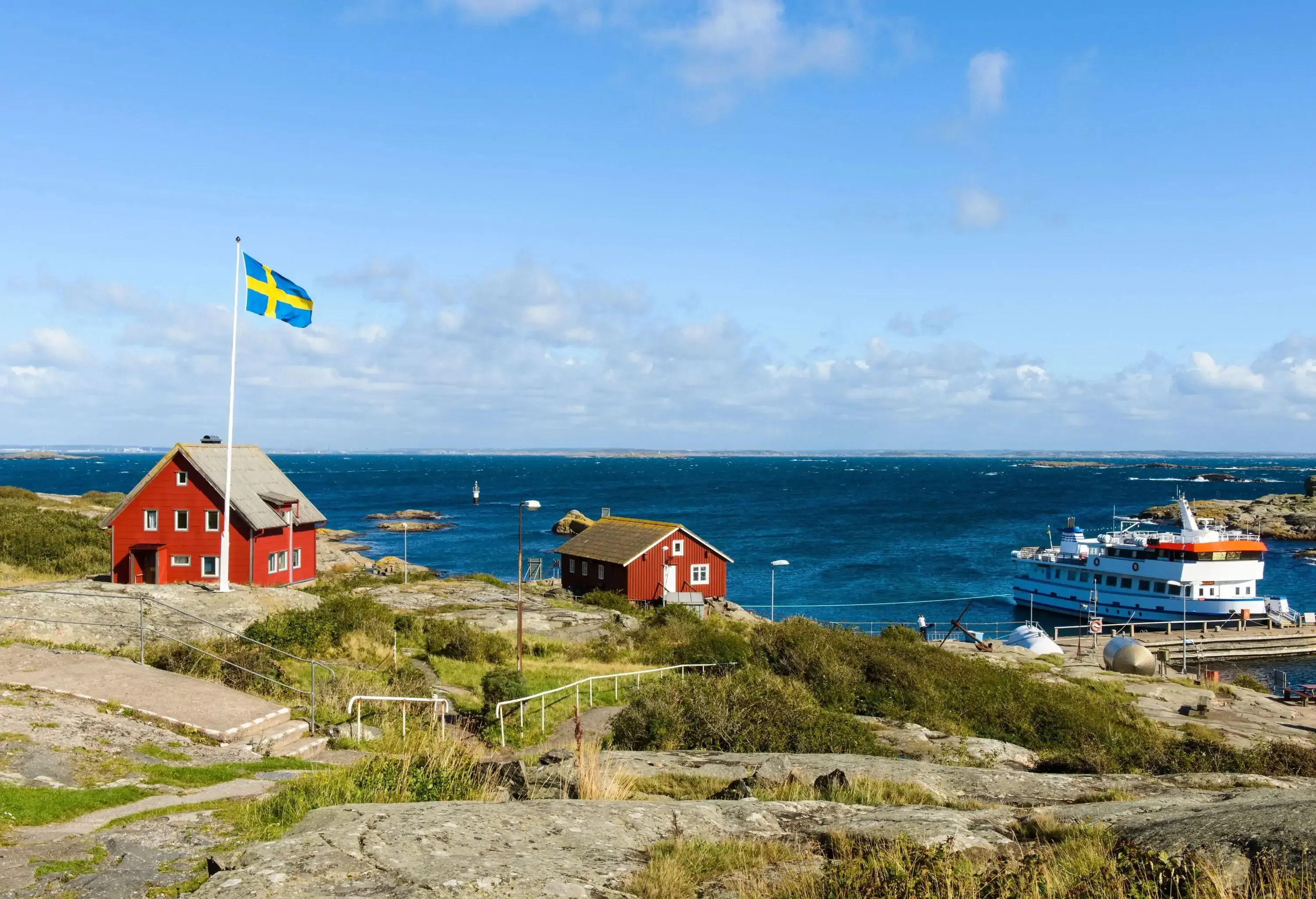 Two maroon cottages and a Swedish flag standing on the rocky coast with a docked ferry boat.