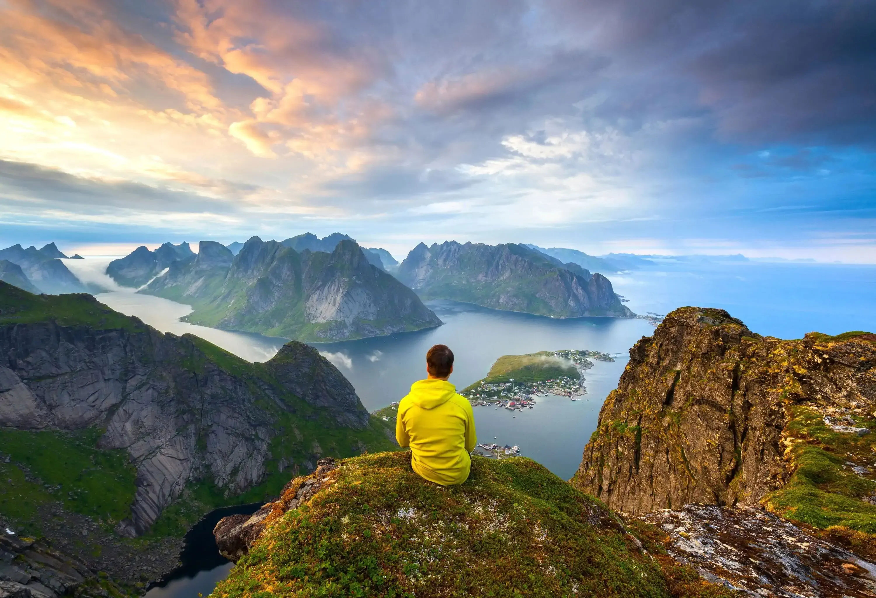 A man sits atop a rocky mountain with a spectacular view of steep rock islands emerging on the sea against the scenic cloudy sky.