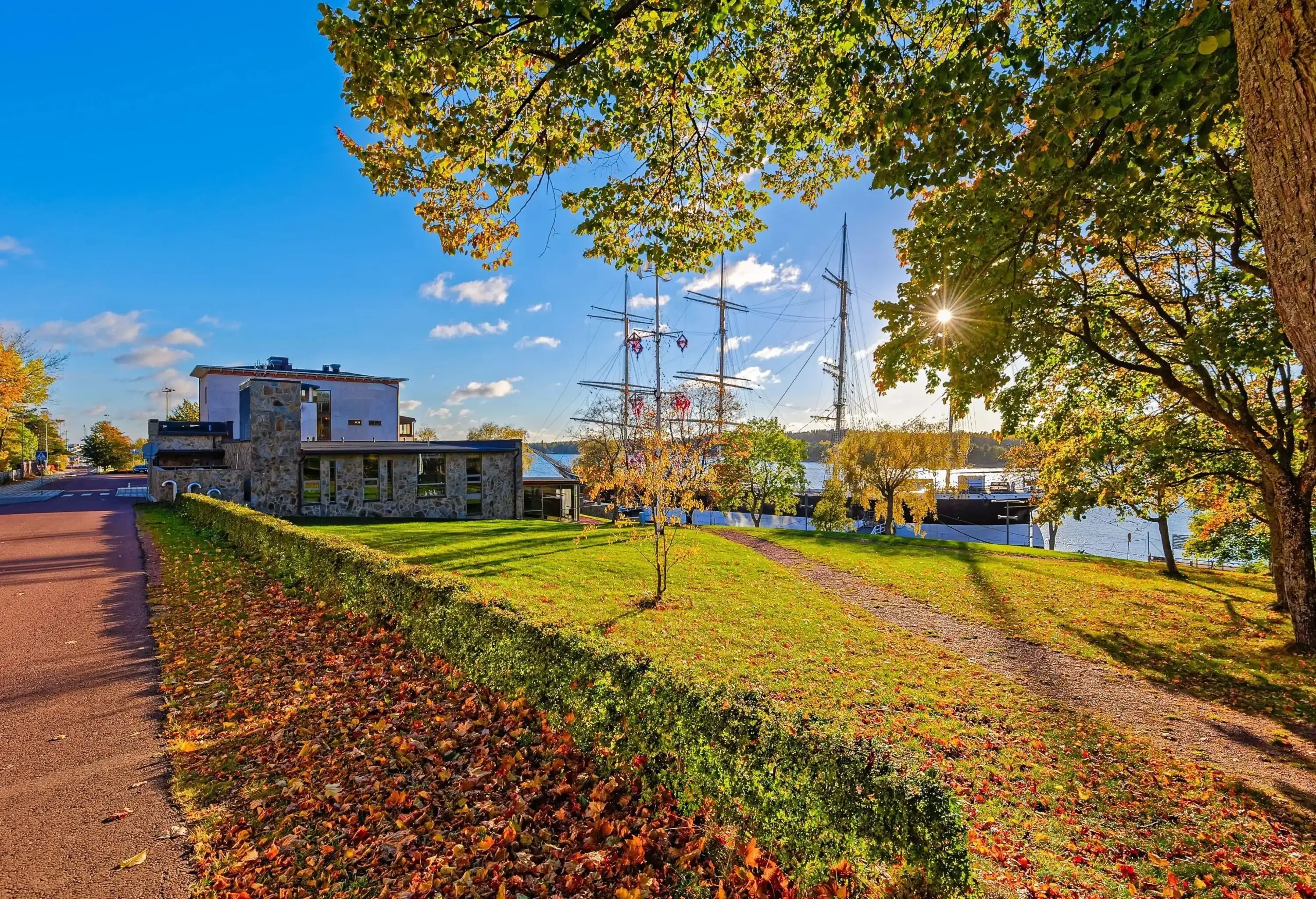 The sun shining through the tree branches on a lawn of a stone house, with an anchored boat on the harbour.