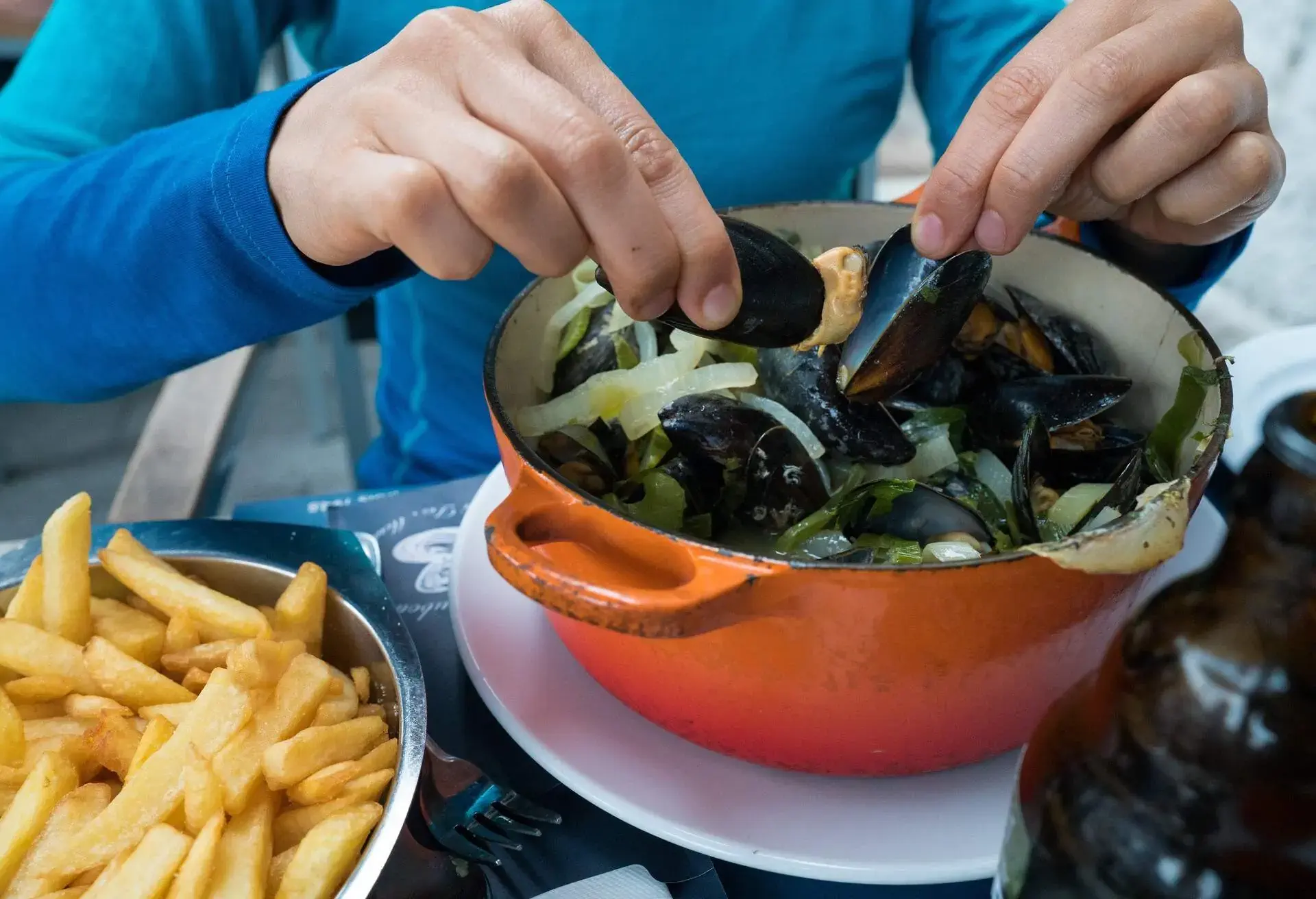 Person in a blue shirt scooping mussels from an orange pot with french fries on the side