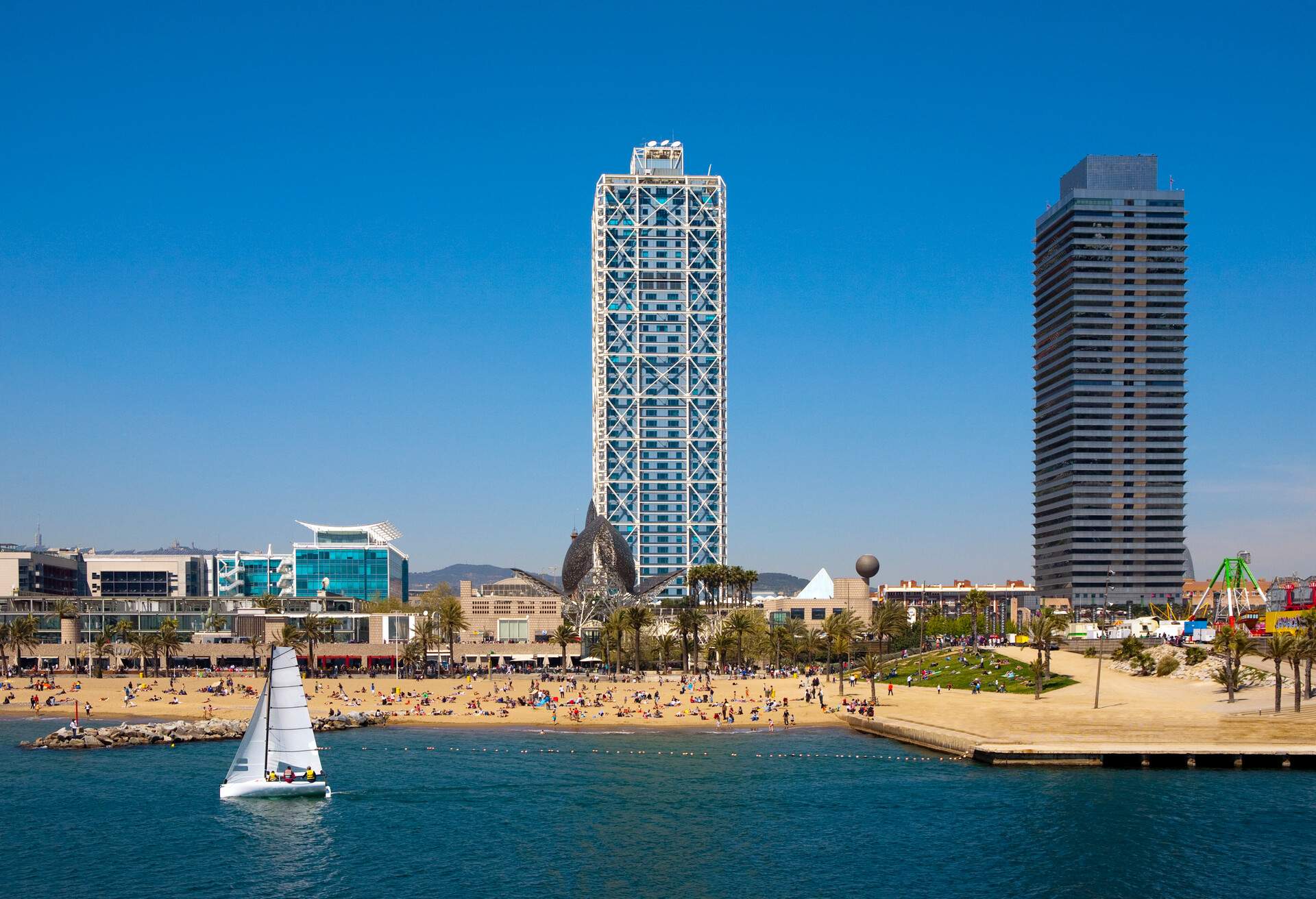 Two tall skyscrapers and other structures along the coast of a populated beach.