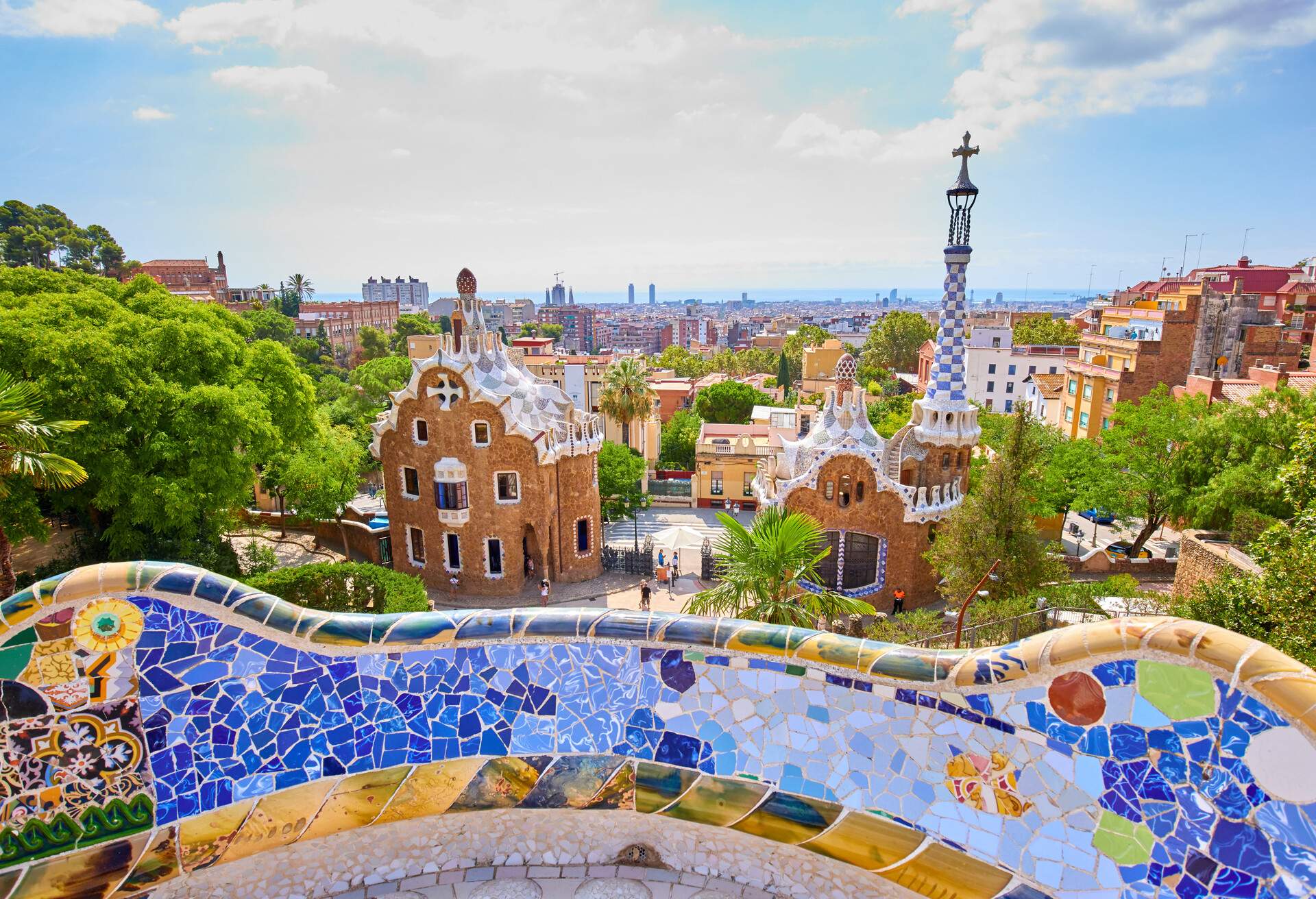 A balcony adorned with a mosaic of colourful tiles overlooks Park Güell, a park with uniquely-designed structures surrounded by trees.