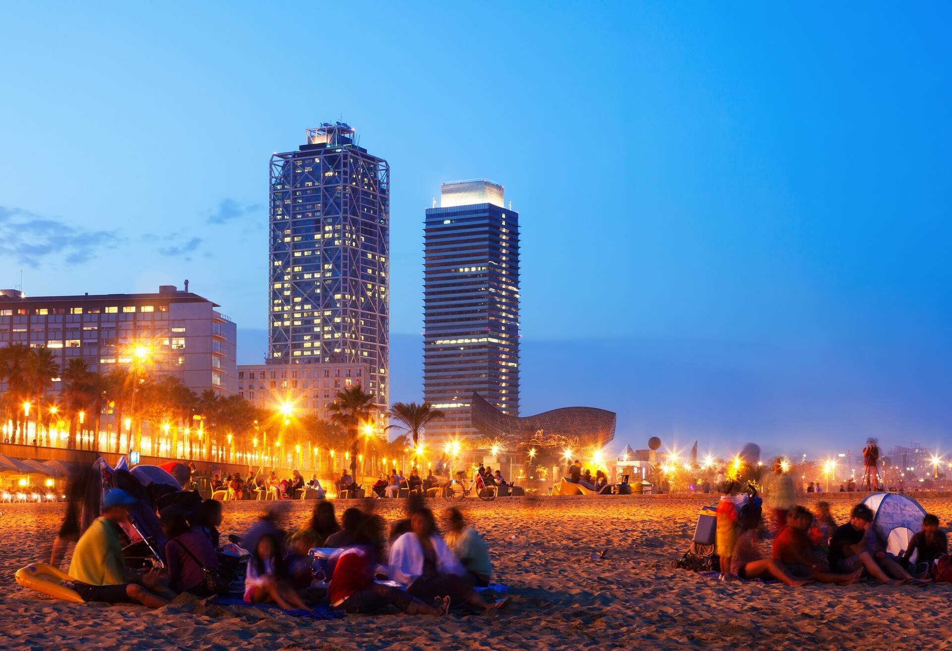 People gather on a beach lined with city lights, with views of two skyscrapers in the background.