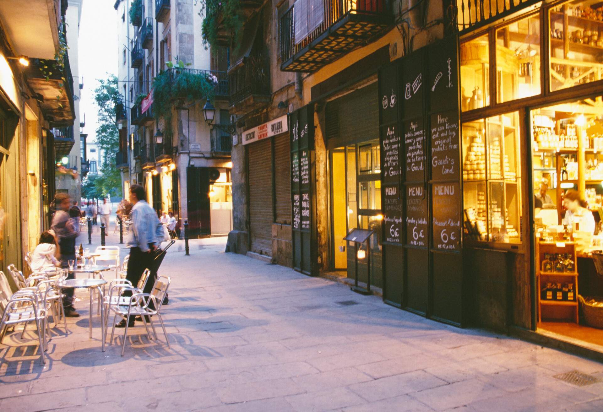 People having drinks on outdoor chairs and tables along a street flanked by illuminated stores.