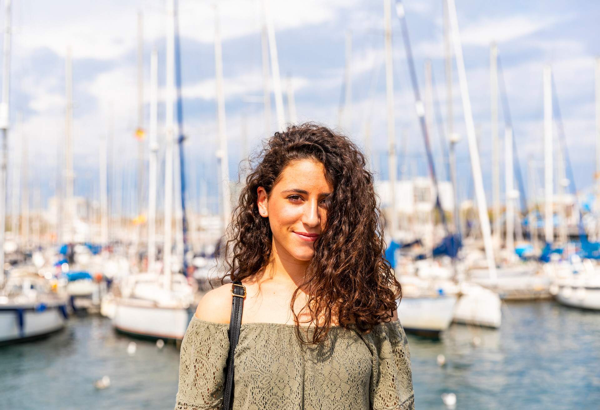 A curly-haired woman smiles against the backdrop of yachts docked on a harbour.