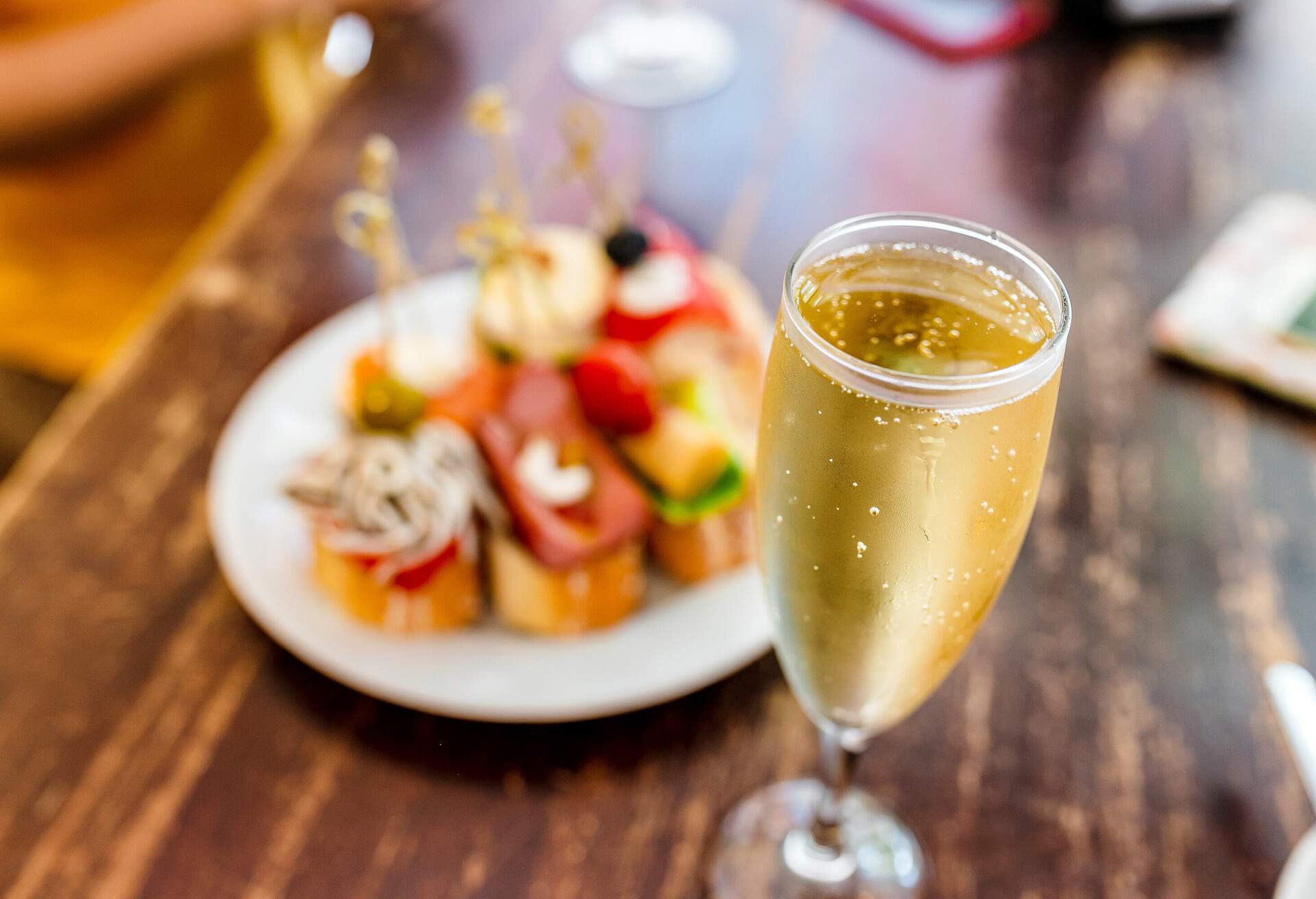 A sparkling glass of Cava wine takes centre stage in the foreground, while a sumptuous plate of food sits tantalisingly in the softly blurred background.